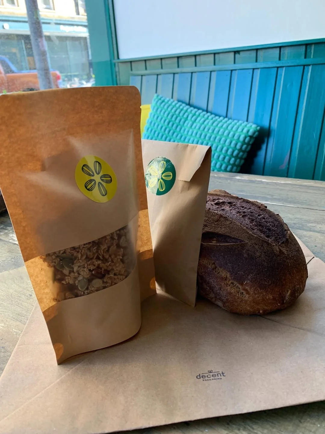 Two brown paper bags with yellow and green circular logos, a loaf of gluten-free rustic bread, on a tray with the word 'decent' printed on it, inside a restaurant with a teal wood-paneled wall and a teal textured pillow on a booth.