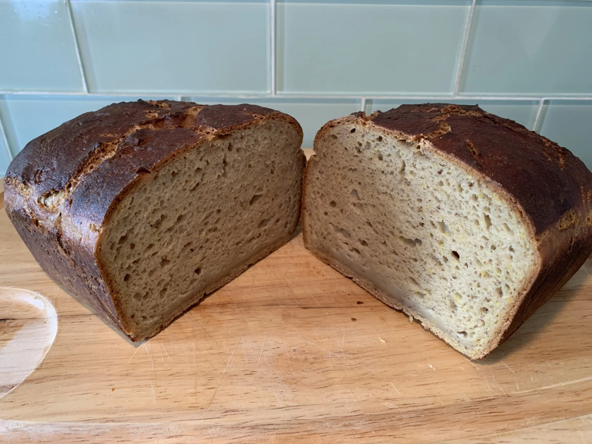 A freshly baked loaf of gluten-free bread sliced in half on a wooden cutting board, with a small dish of butter nearby, in a kitchen setting with a tiled backsplash.