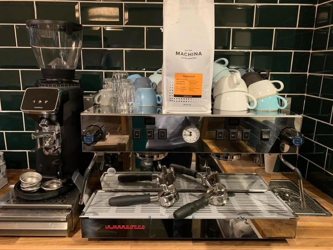 A coffee station with an espresso machine, cups, glasses, coffee beans, and a coffee grinder against a dark green tiled wall.