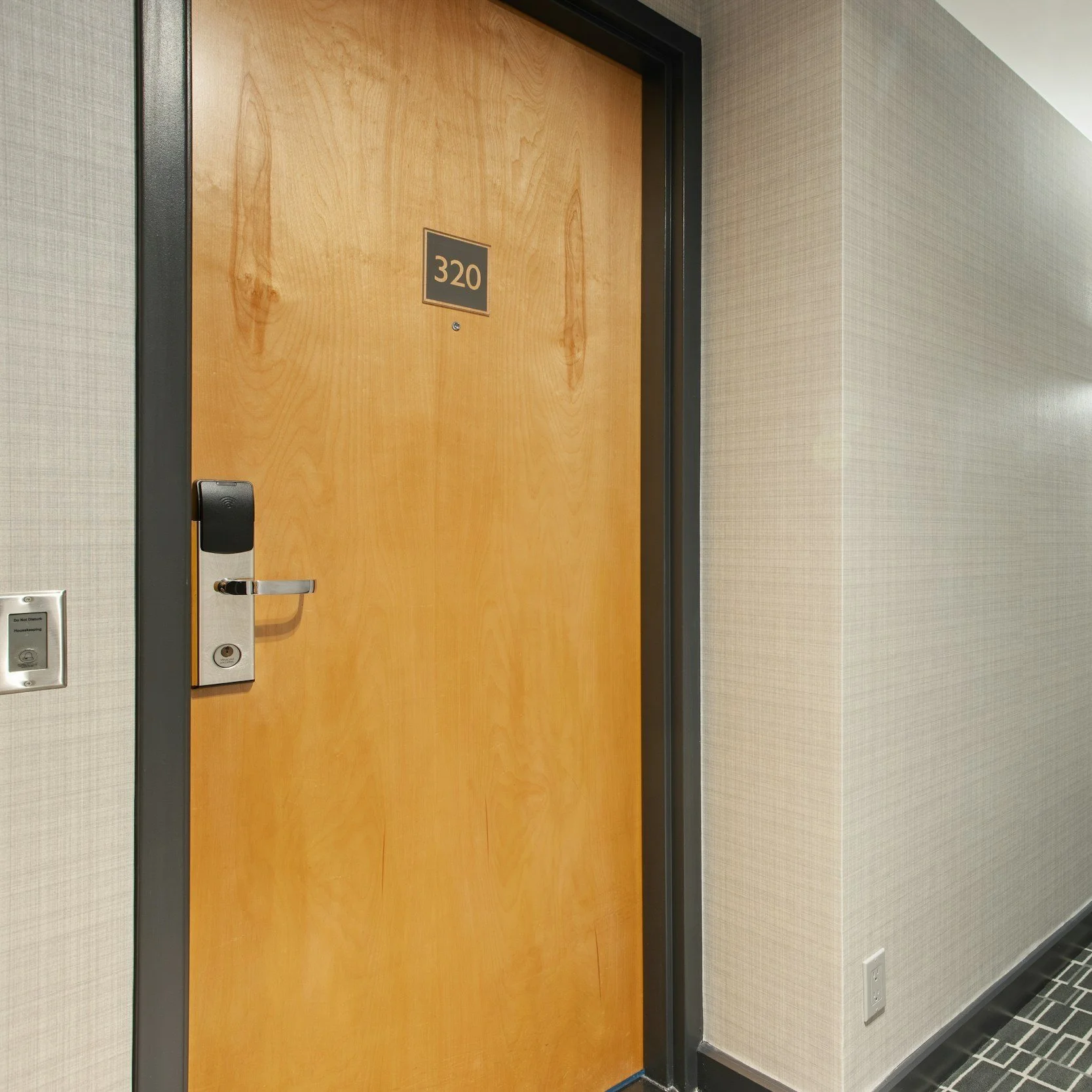 Hotel interior hallway with a wooden door labeled 320, electronic lock, beige textured walls, and a power outlet near the floor.