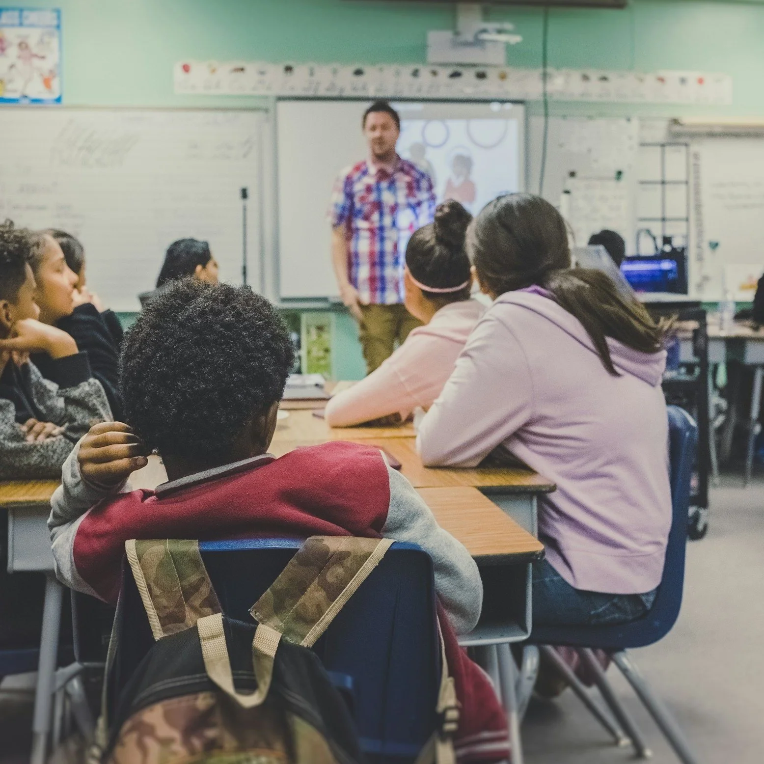 Students sitting at desks in a classroom listening to a teacher standing in front of a whiteboard.