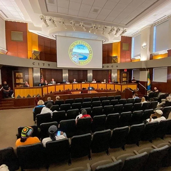 Municipal meeting room with a curved set of desks and several officials seated behind them. There is an audience watching, with some people wearing hats. A large screen displays the seal of Chattanooga, Tennessee, and the word 'CITY' is visible on the wall behind the officials.