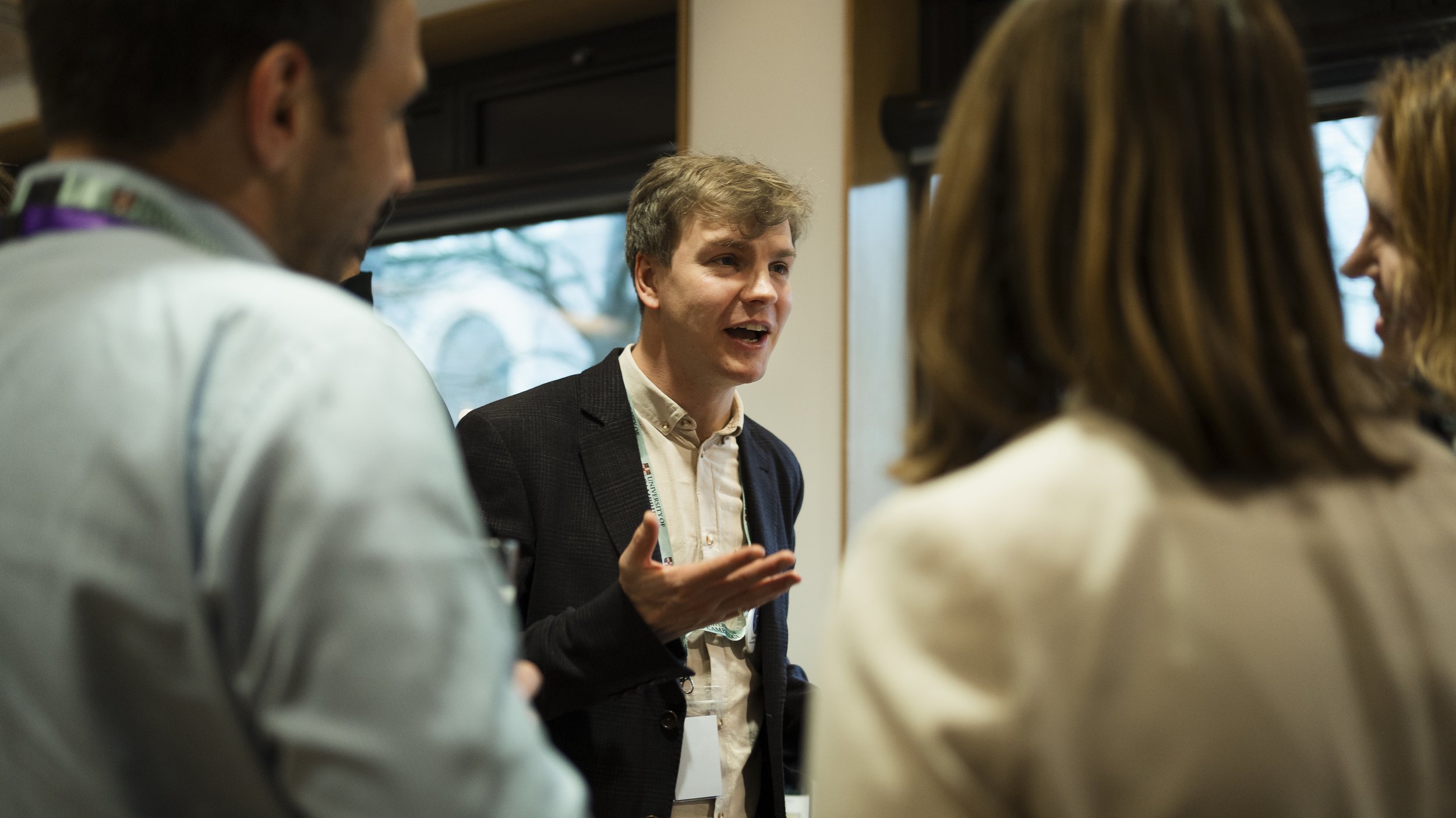A man with curly hair and a dark blazer is speaking to a group of people in a conference room. Several others are visible from behind, listening attentively. Windows with natural light are in the background.