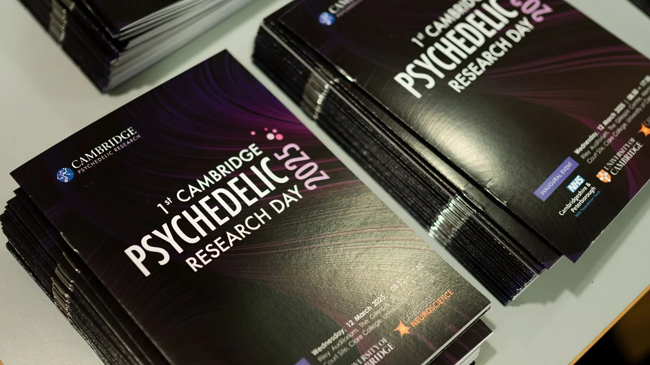 Stacks of brochures for the 1st Cambridge Psychedelic Research Day on March 12, 2023, with a dark purple and black design and logos for Cambridge Psychedelic Research, NHS, UCL, and other organizations.
