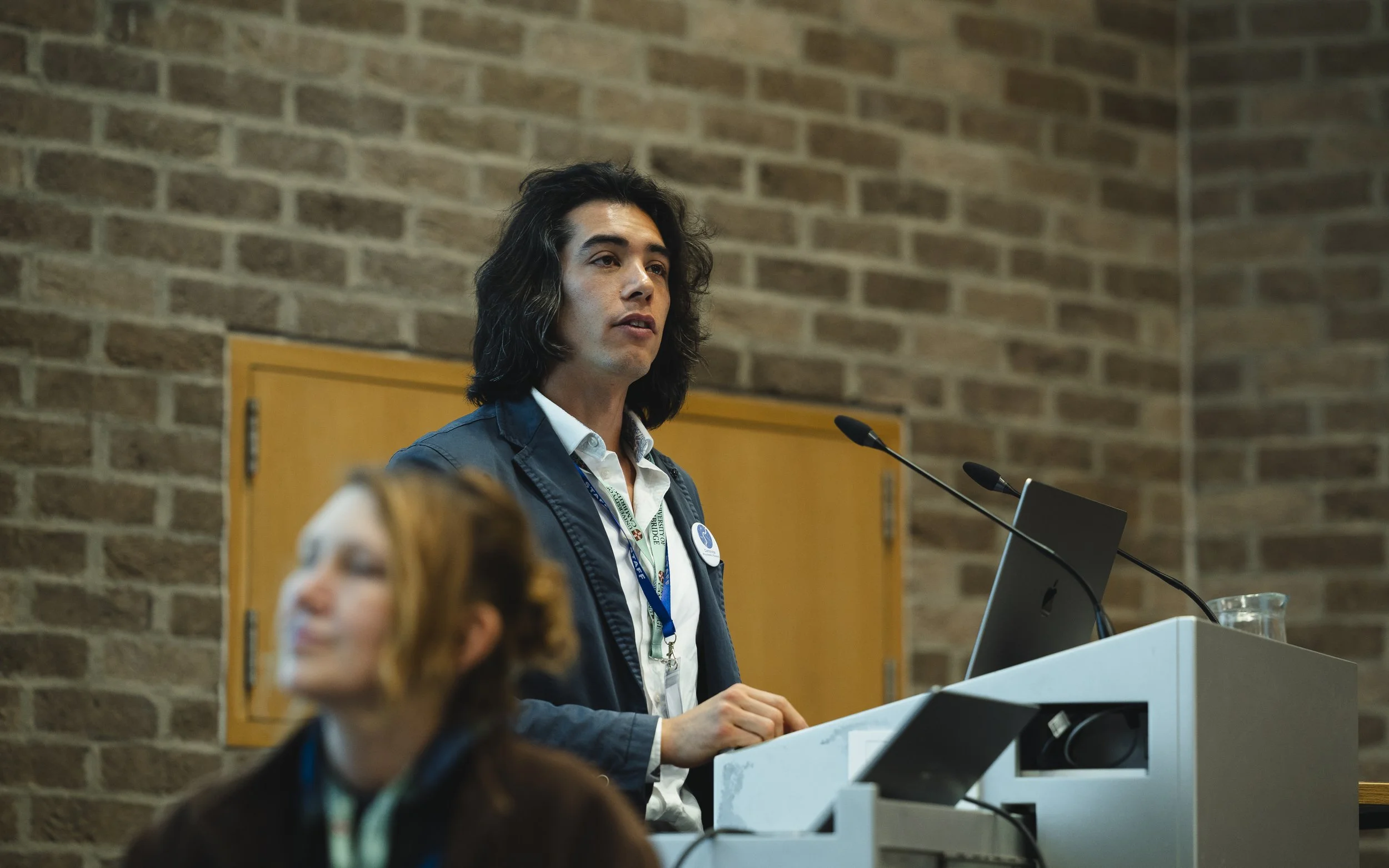 A woman with shoulder-length dark hair, wearing a white shirt and a blue jacket, stands at a podium with microphones, speaking in front of a brick wall during a conference or presentation.