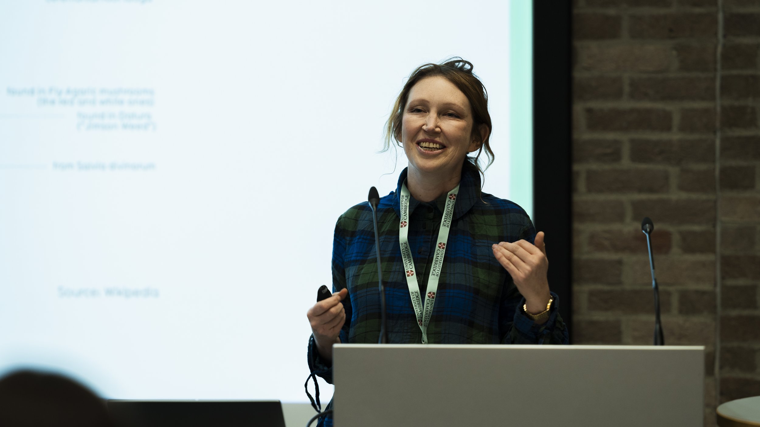 A woman with brown hair wearing a blue and green plaid shirt, standing at a podium, smiling and gesturing with her right hand during a presentation. She has a university lanyard around her neck and a brick wall in the background.