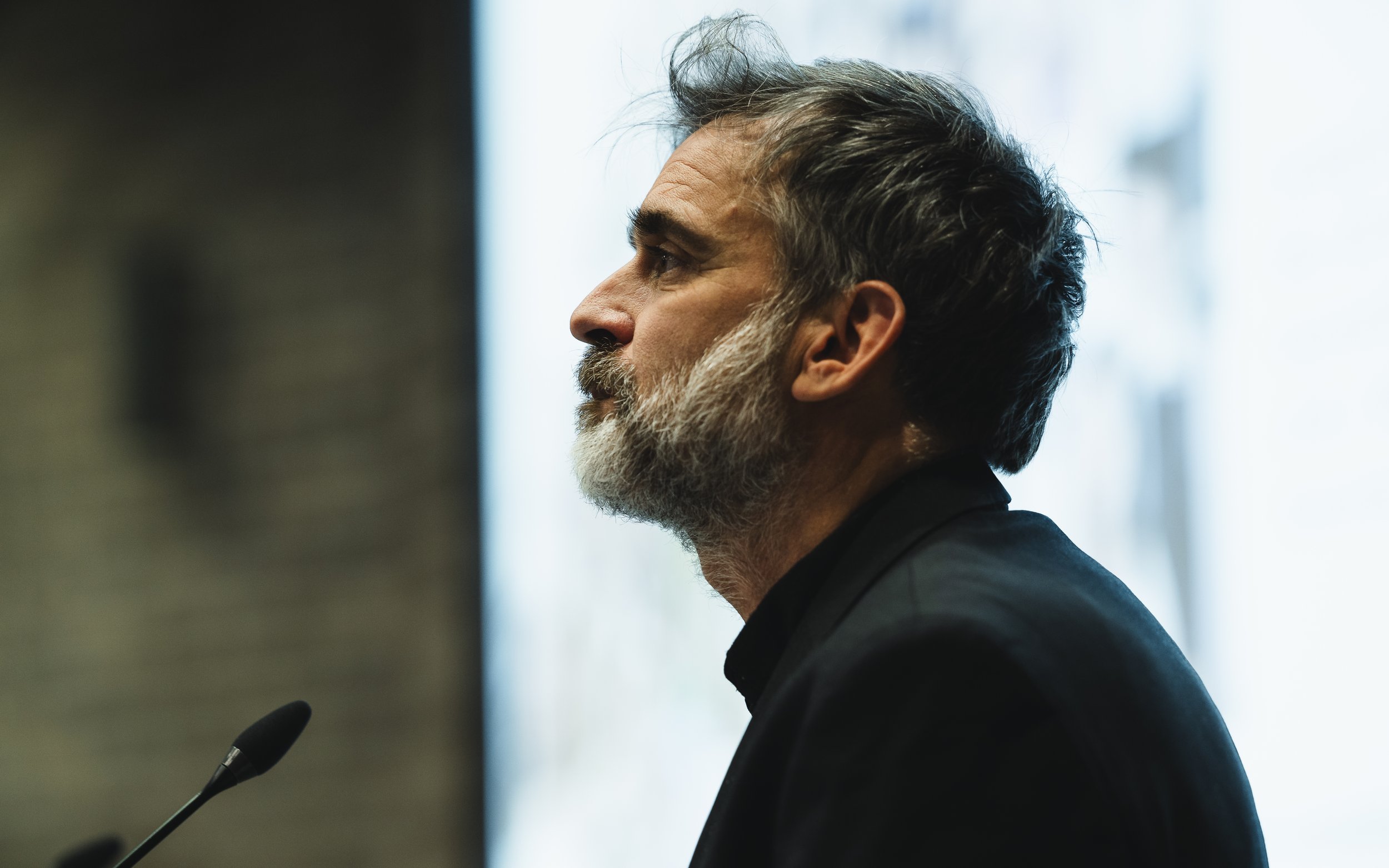 Side profile of a middle-aged man with gray hair and a beard, wearing a dark suit, sitting in front of a bright window.