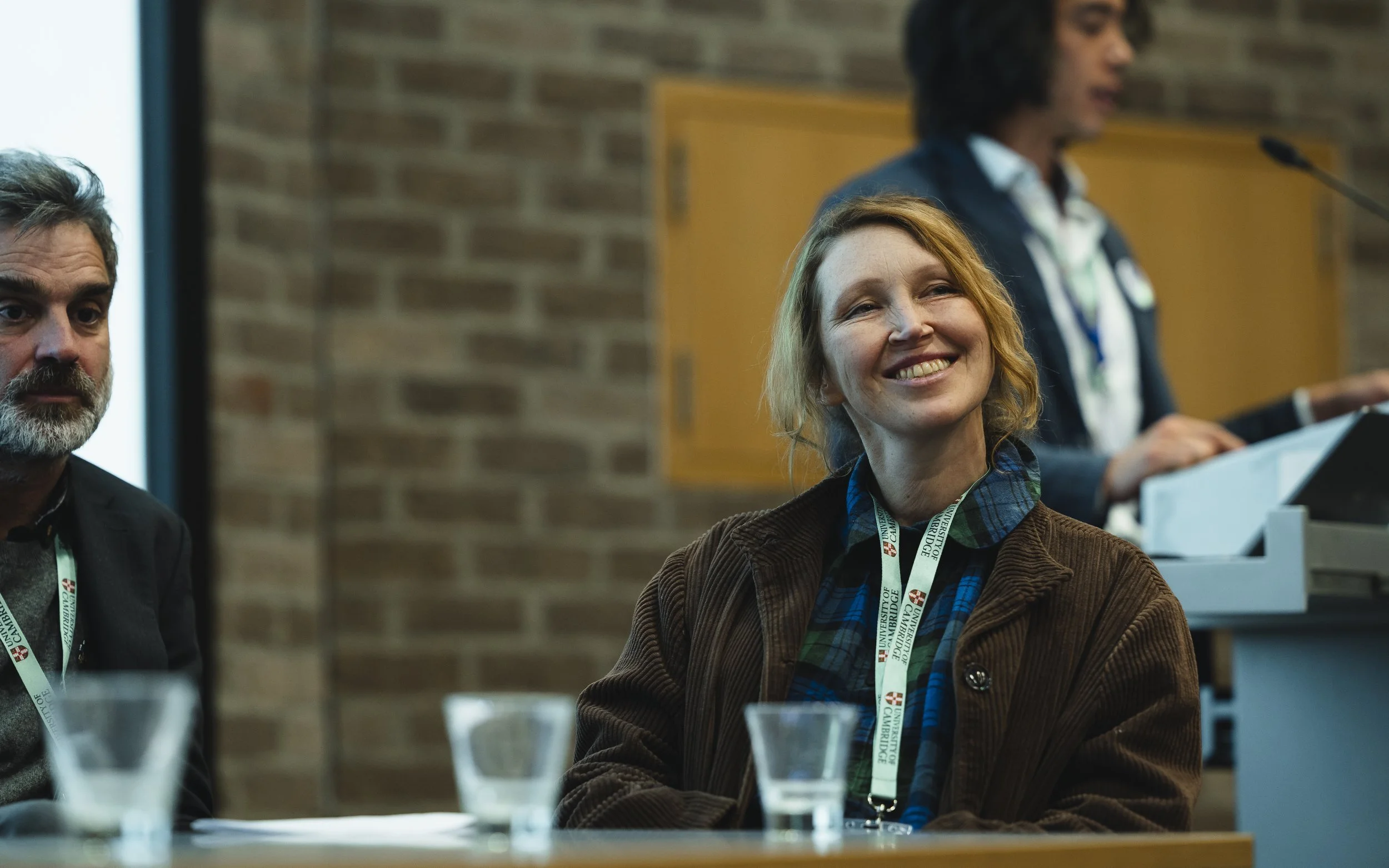 A woman with red hair and a brown jacket smiling at a conference or meeting, with a man and another woman visible in the background. The woman is wearing a lanyard from the University of Cambridge.