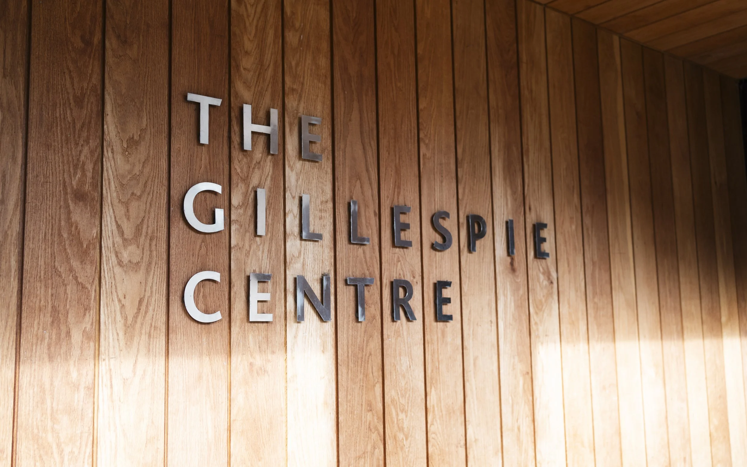 Wood-paneled wall with metal letters spelling 'THE GILLESPIE CENTRE' mounted on it.