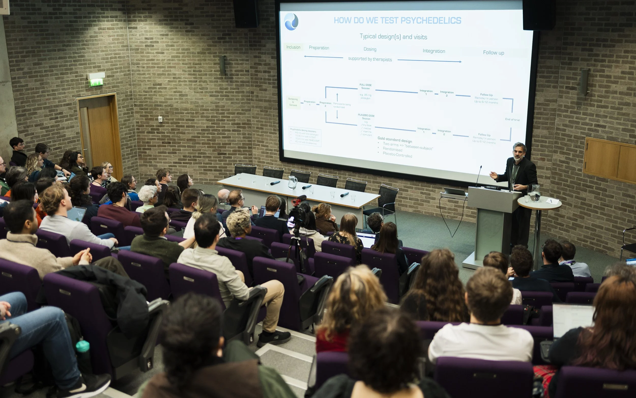 A scientist giving a presentation on testing psychedelics in a lecture hall filled with an audience.