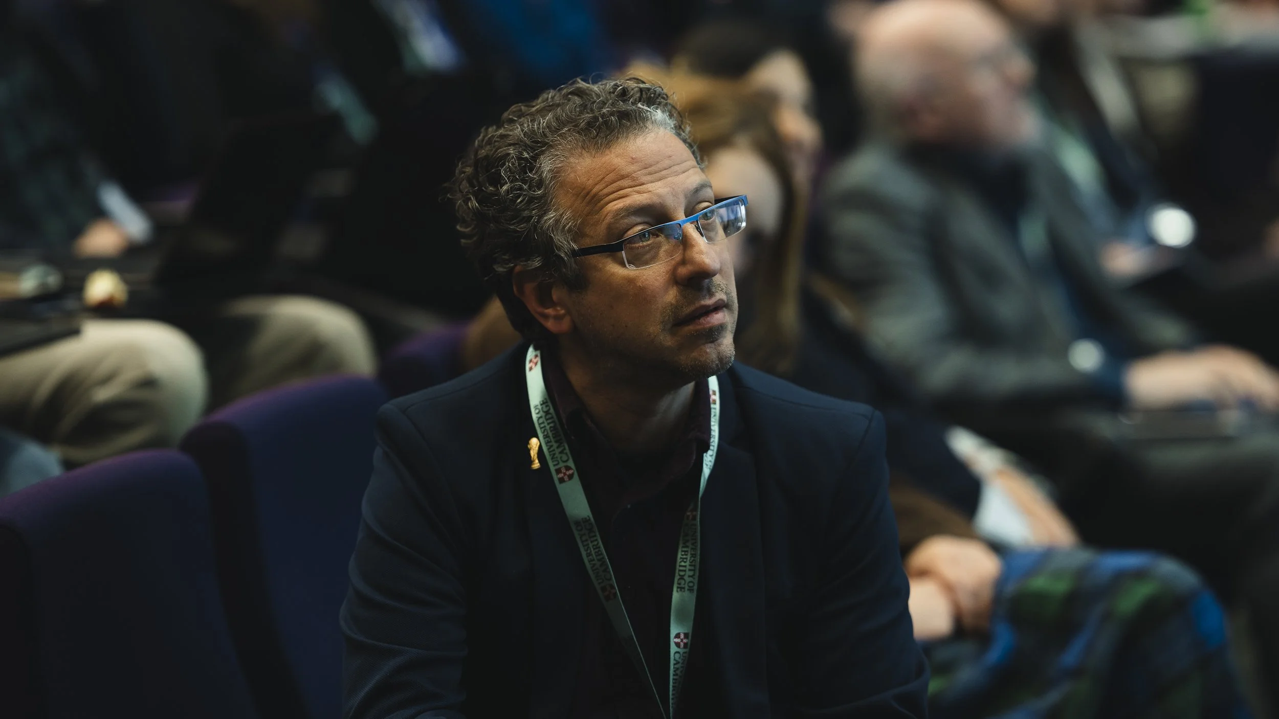 A man with curly gray hair and glasses sitting in an audience at a conference, wearing a black jacket and a conference badge around his neck.
