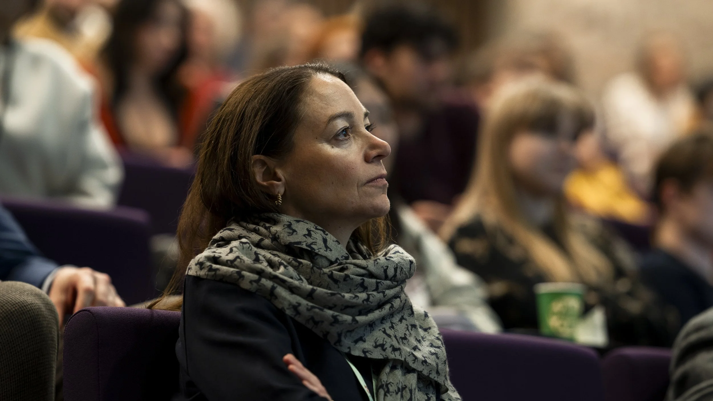 A woman with brown hair wearing a black jacket and a gray scarf with black dog patterns, attentively listening at a conference or seminar surrounded by other attendees.