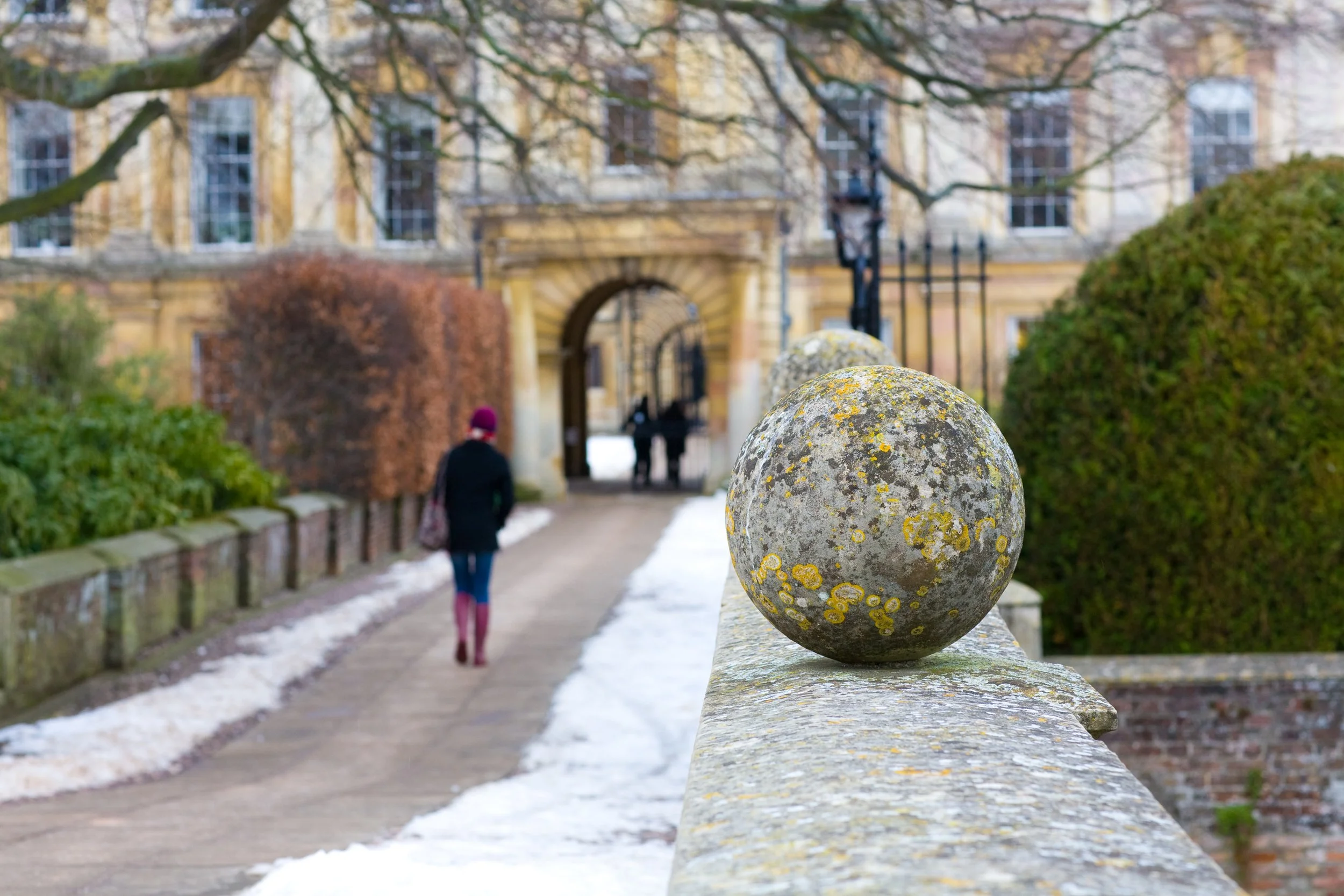 Close-up of a lichen-covered stone ball on a stone fence with a blurred background of a university or historical building, bushes, and a person walking on a snowy pathway.