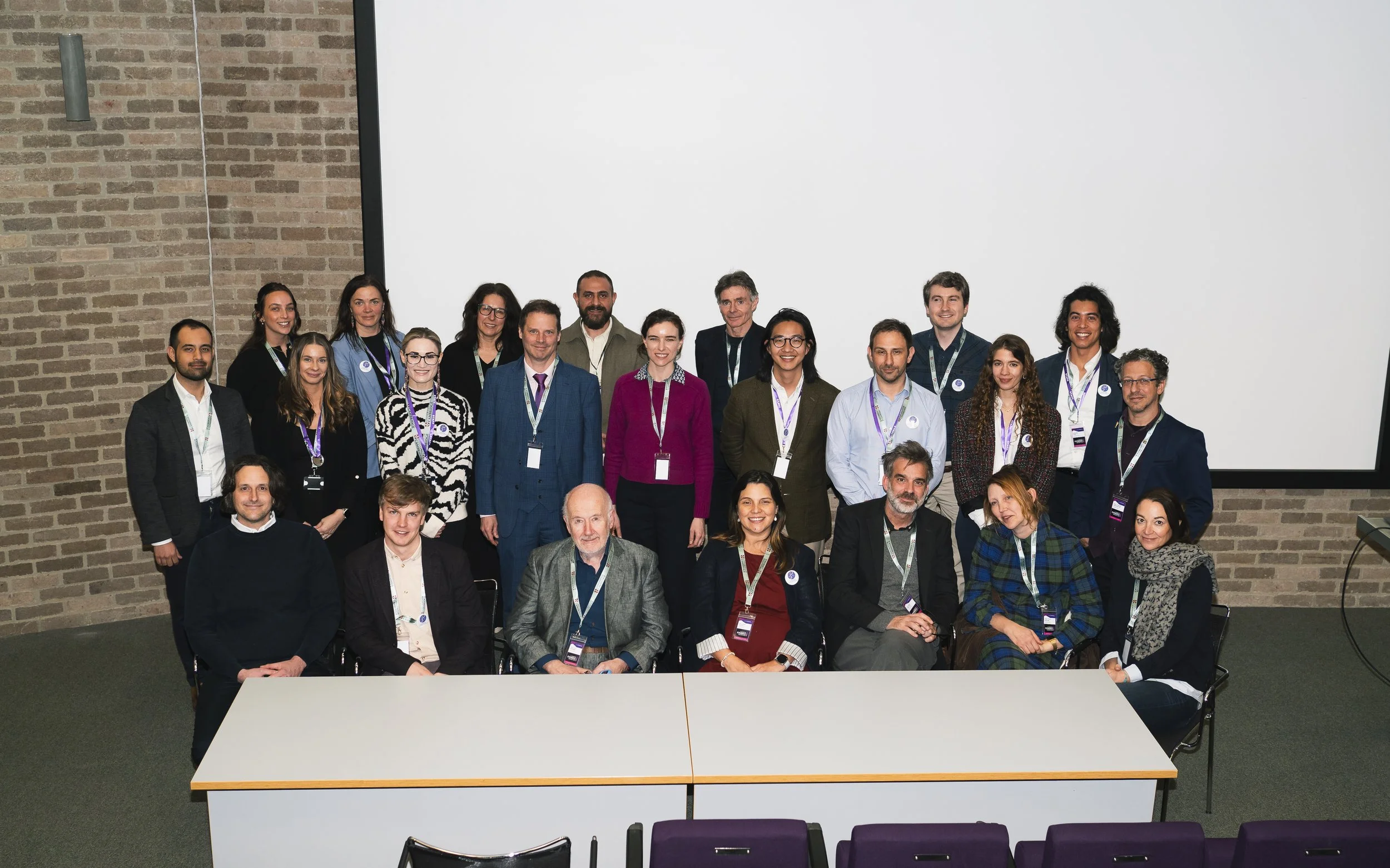 A large group of people, including men and women of various ages, gathered for a group photo in a conference or event setting. They are standing and sitting in front of a large white screen with a brick wall to the left. Many are wearing conference b