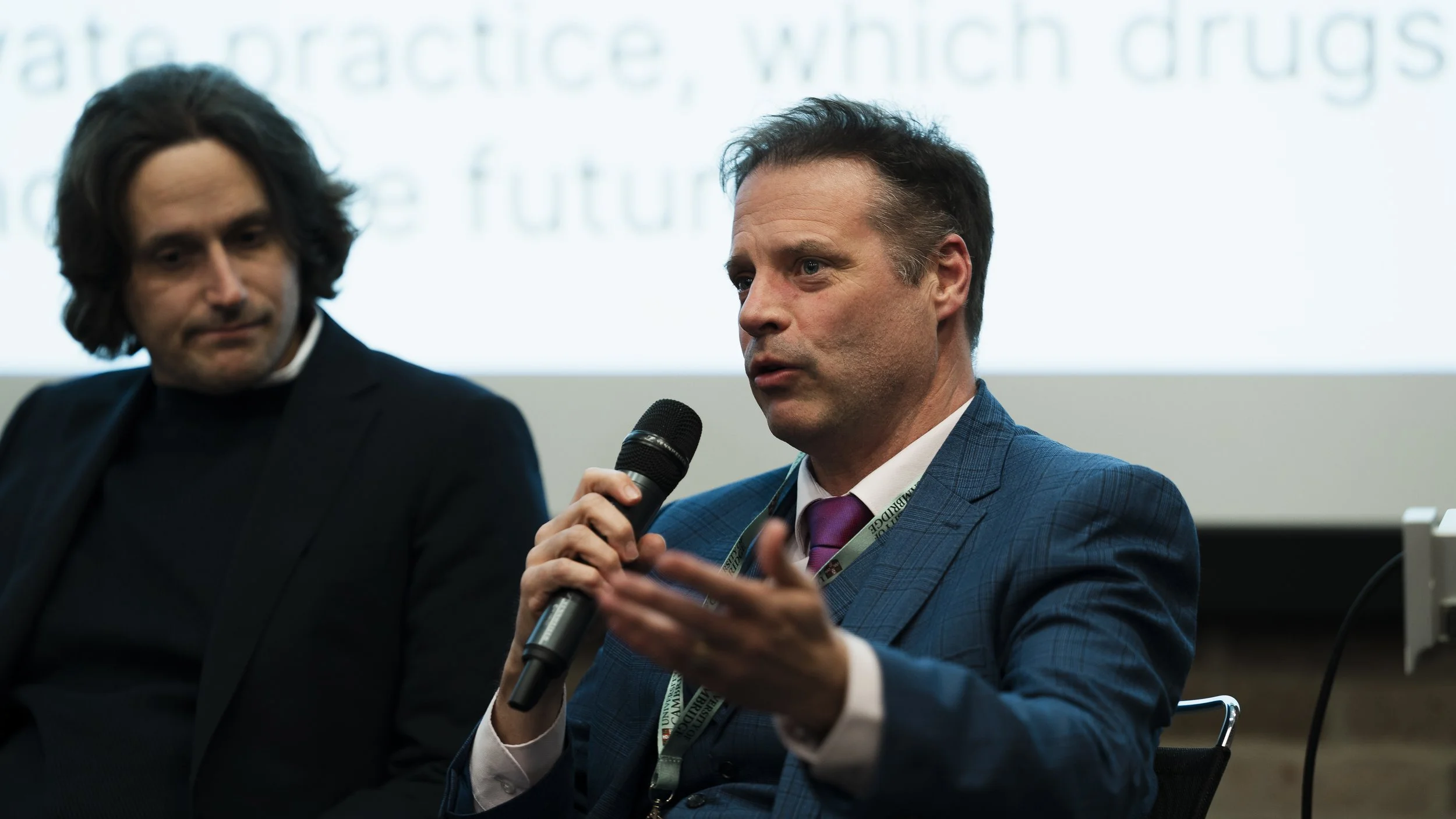 Two men speaking at a conference or panel discussion, one with a microphone and the other looking on, with a blurred background of a presentation screen.