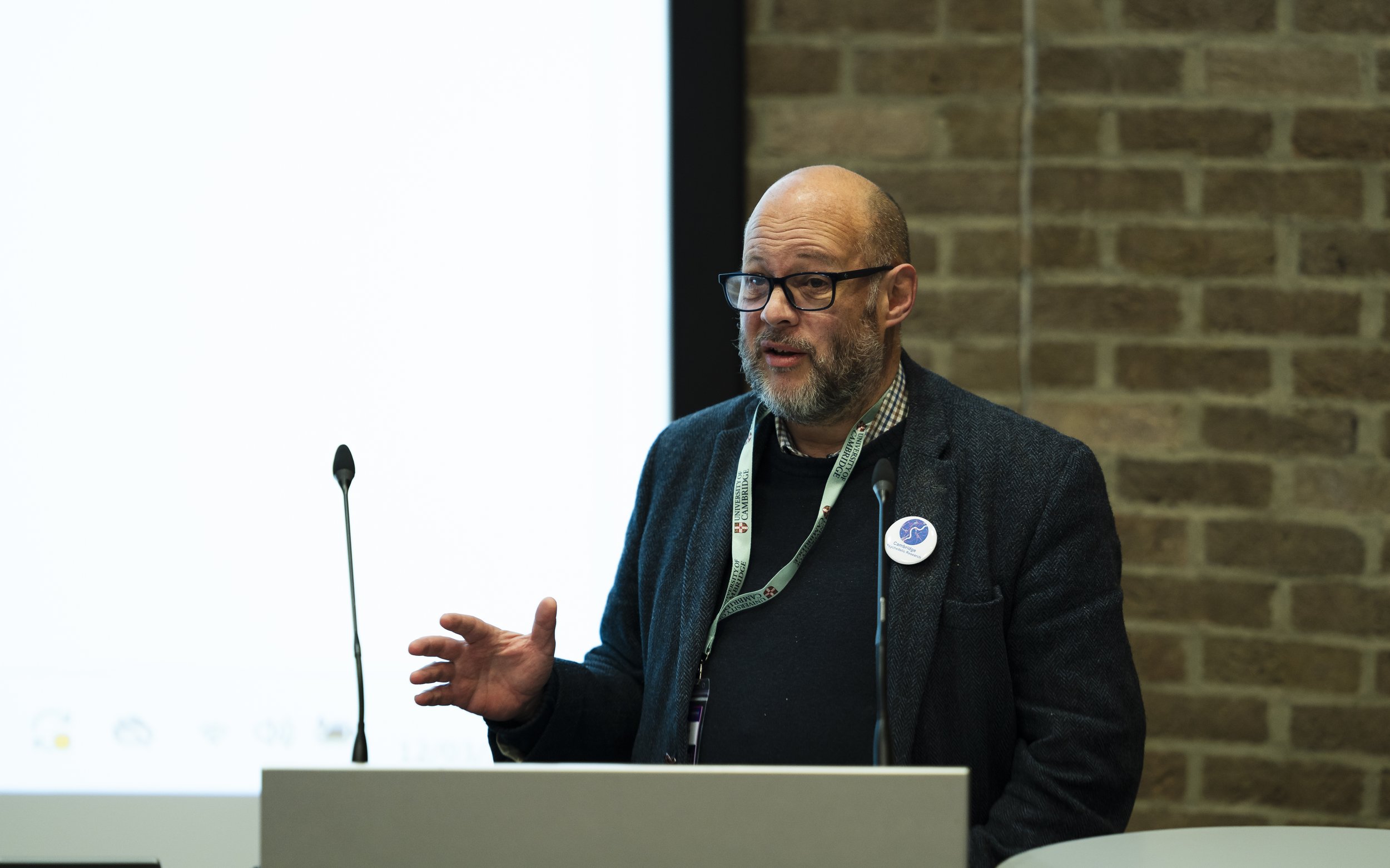 A man with glasses and a beard giving a presentation in front of a brick wall, with a projection screen to his left.