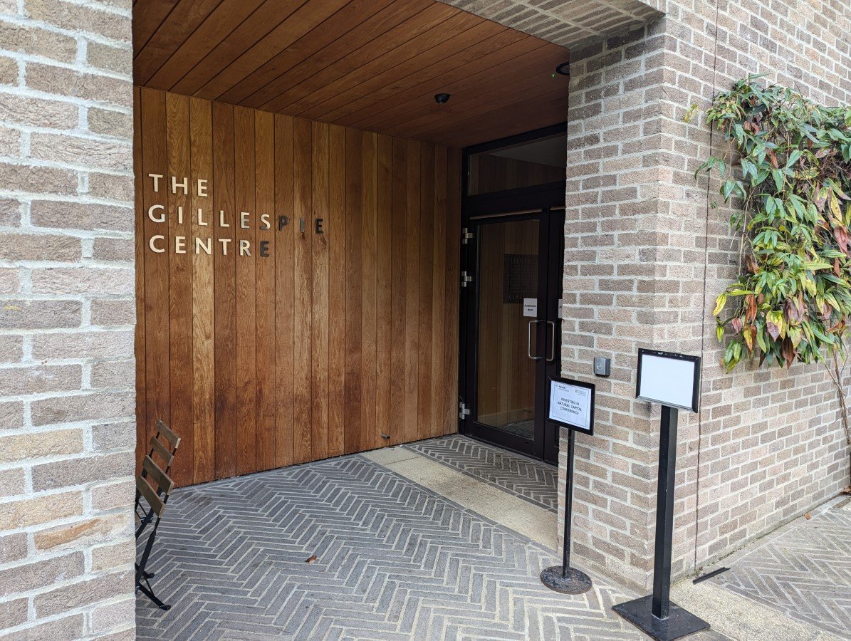 Entrance of The Gillespie Centre with brick walls, a wooden wall, glass door, and signboards.