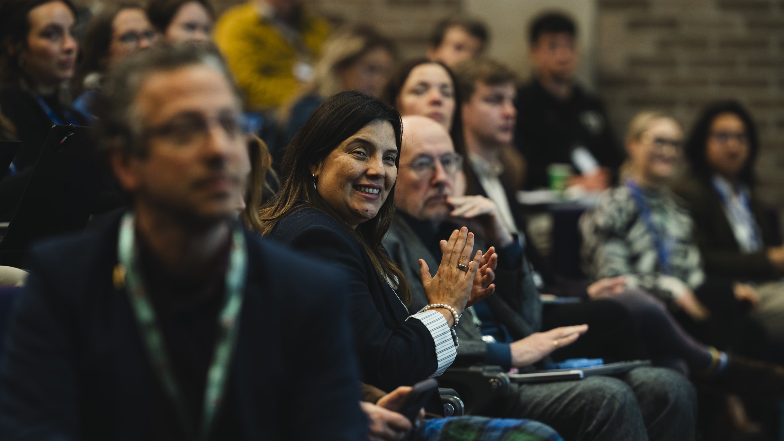 A diverse group of people sitting in an audience at a conference or seminar, listening attentively, with one woman smiling and clapping.
