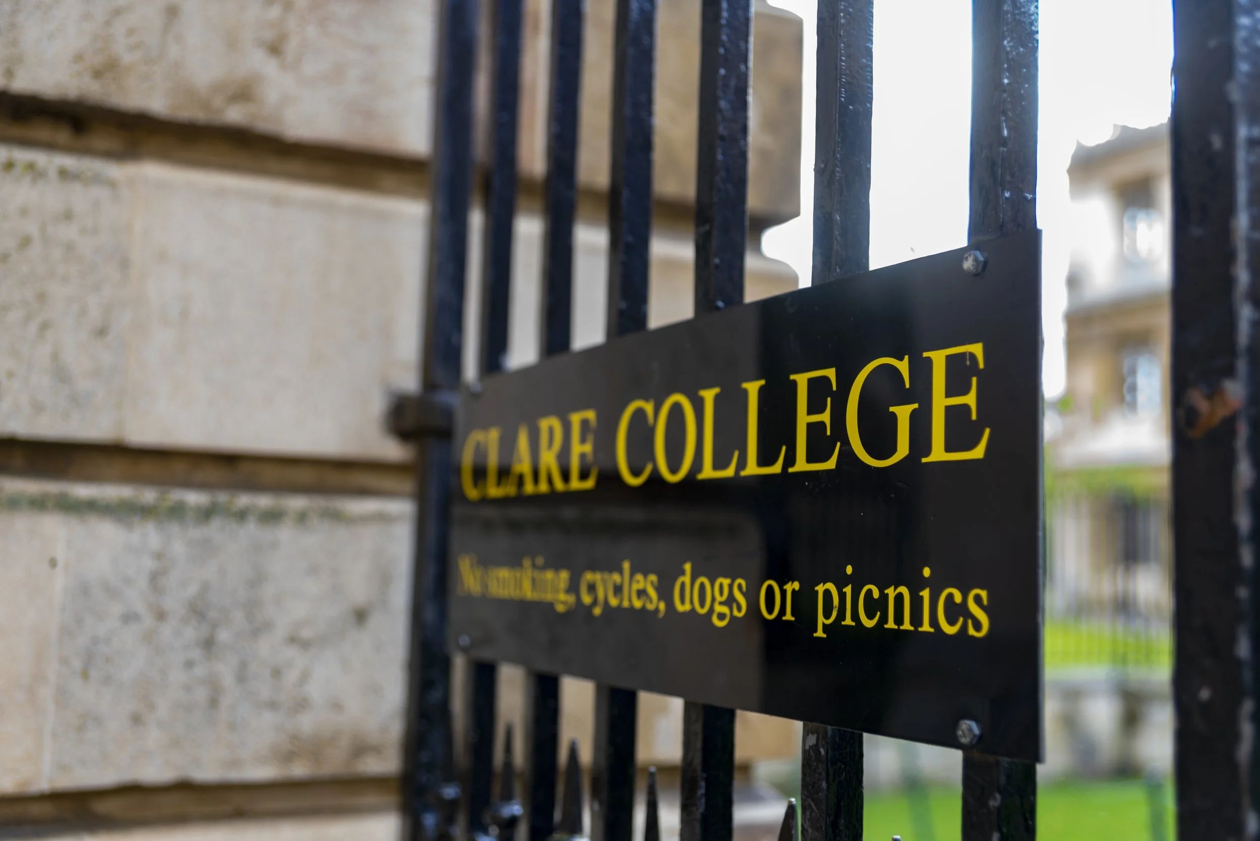 A black metal fence with a yellow sign that reads 'CLARE COLLEGE'. The sign lists restrictions for parking bicycles, dogs, or picnics.