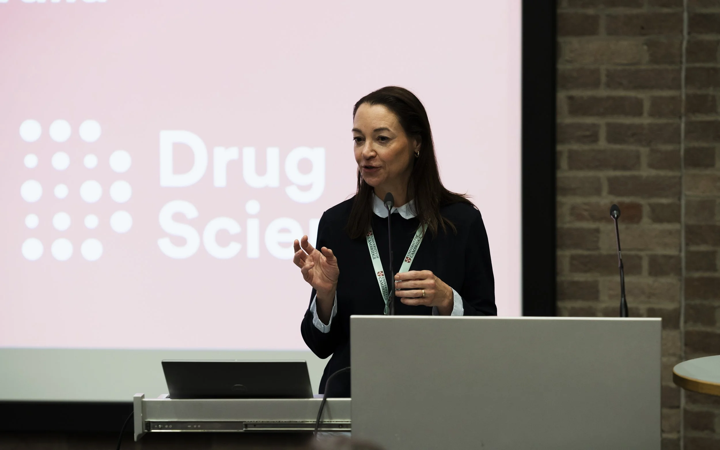 A woman with dark hair and light skin standing behind a podium, giving a presentation. She is gesturing with her right hand and wearing a black blazer over a white shirt. There is a microphone in front of her and a large screen behind her displaying 