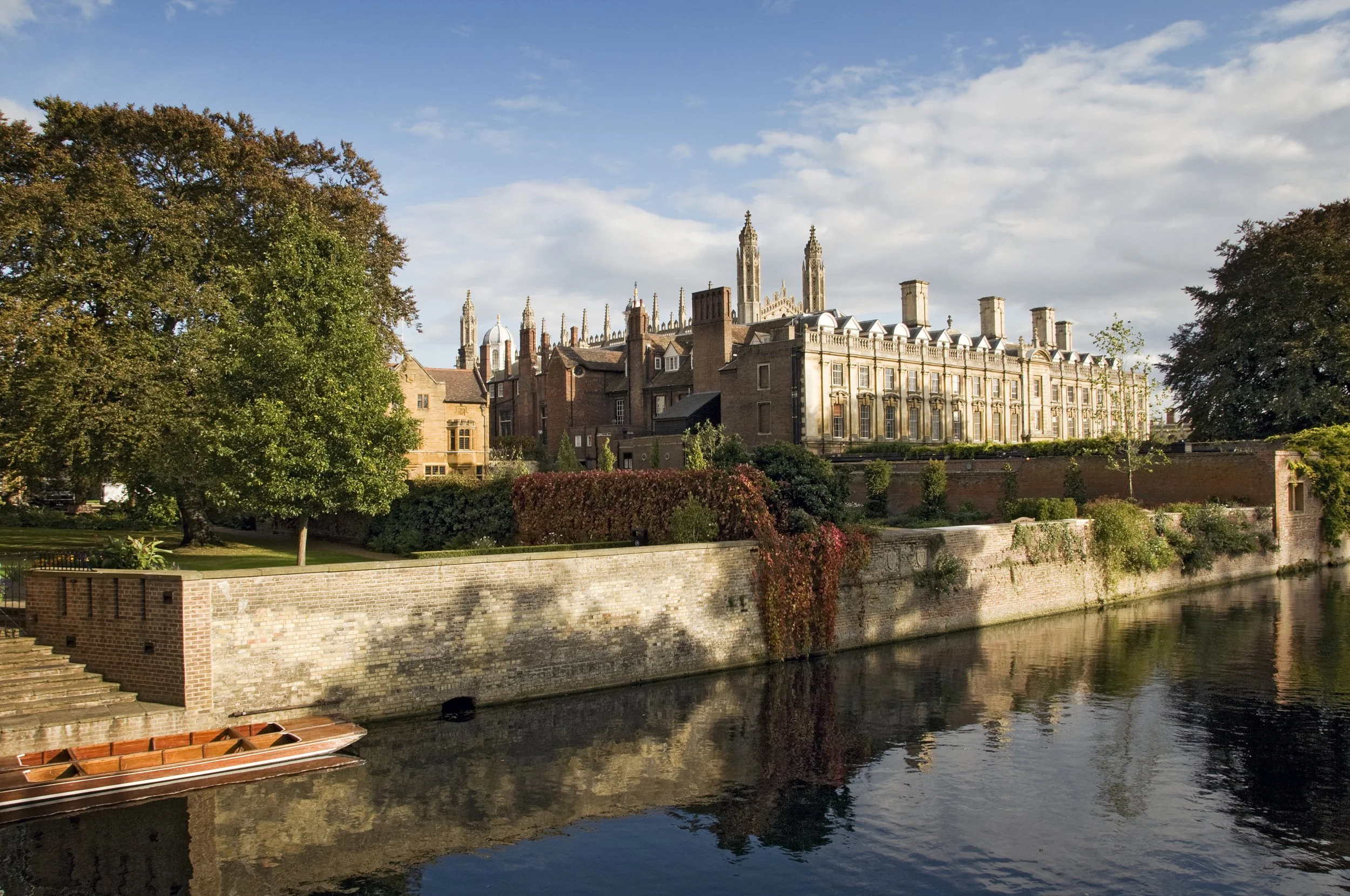 Historic building with Gothic architecture next to a river, surrounded by trees and cloudy sky.