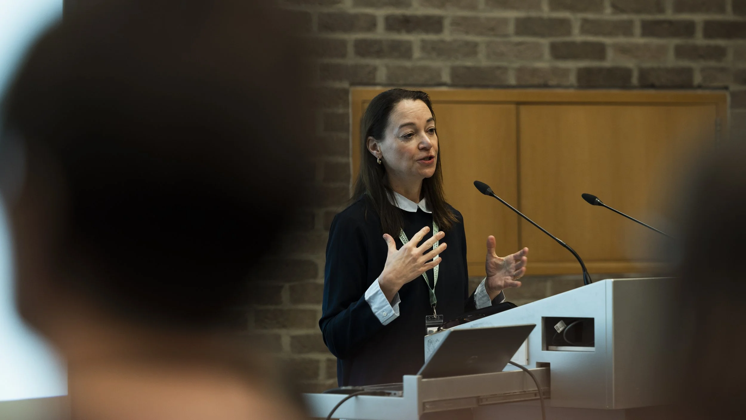 A woman speaking at a podium during a conference or presentation, with a brick wall in the background.