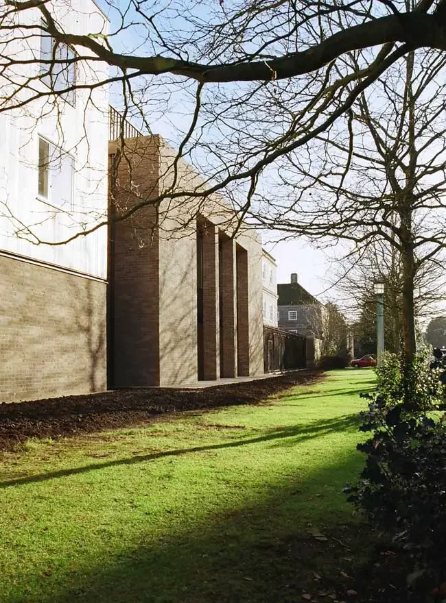 A residential street with a grassy area, trees without leaves, and a building with a brick exterior. The sun is shining, casting shadows of the trees on the ground and building.