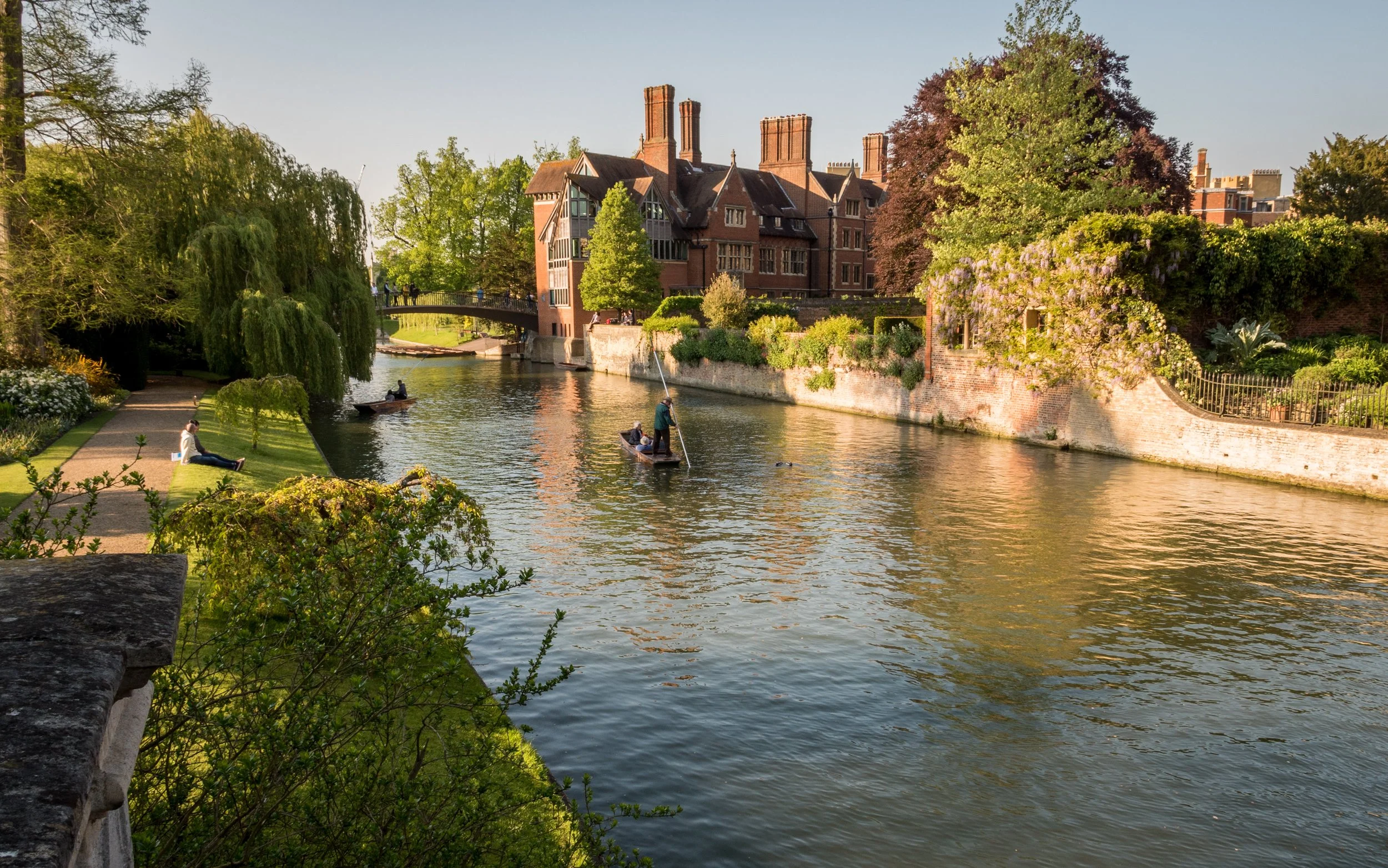 A scenic view of a river with people boating, a walking person, surrounded by green trees, flowering bushes, and a large red brick building with chimneys in the background during daylight.