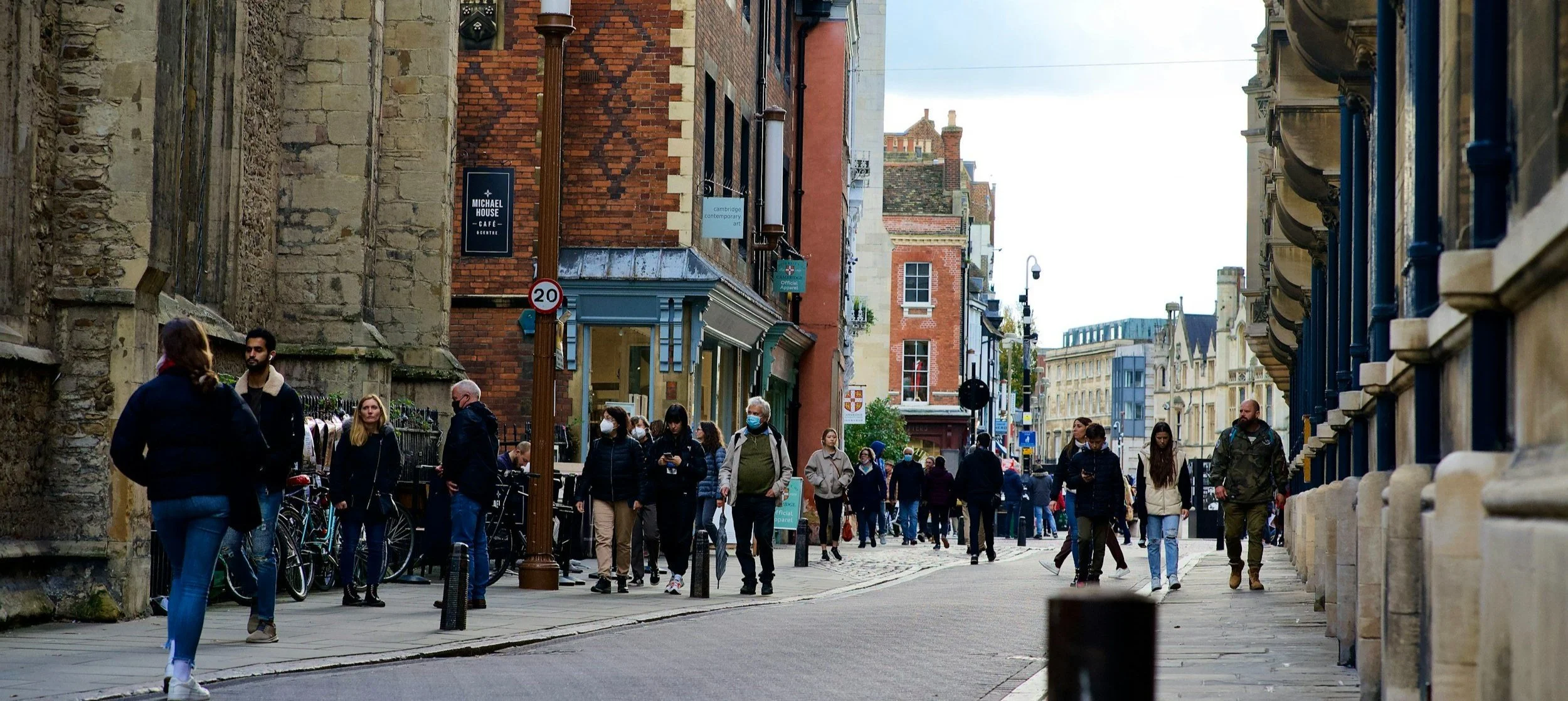 A busy street scene with pedestrians, some wearing face masks, walking along the sidewalk. Bicycles are parked along a fence next to an old stone building, and various shops and buildings line the street. A street sign indicates a speed limit of 20, and there are several street lamps and banners hanging from poles. The weather appears to be cool and overcast.