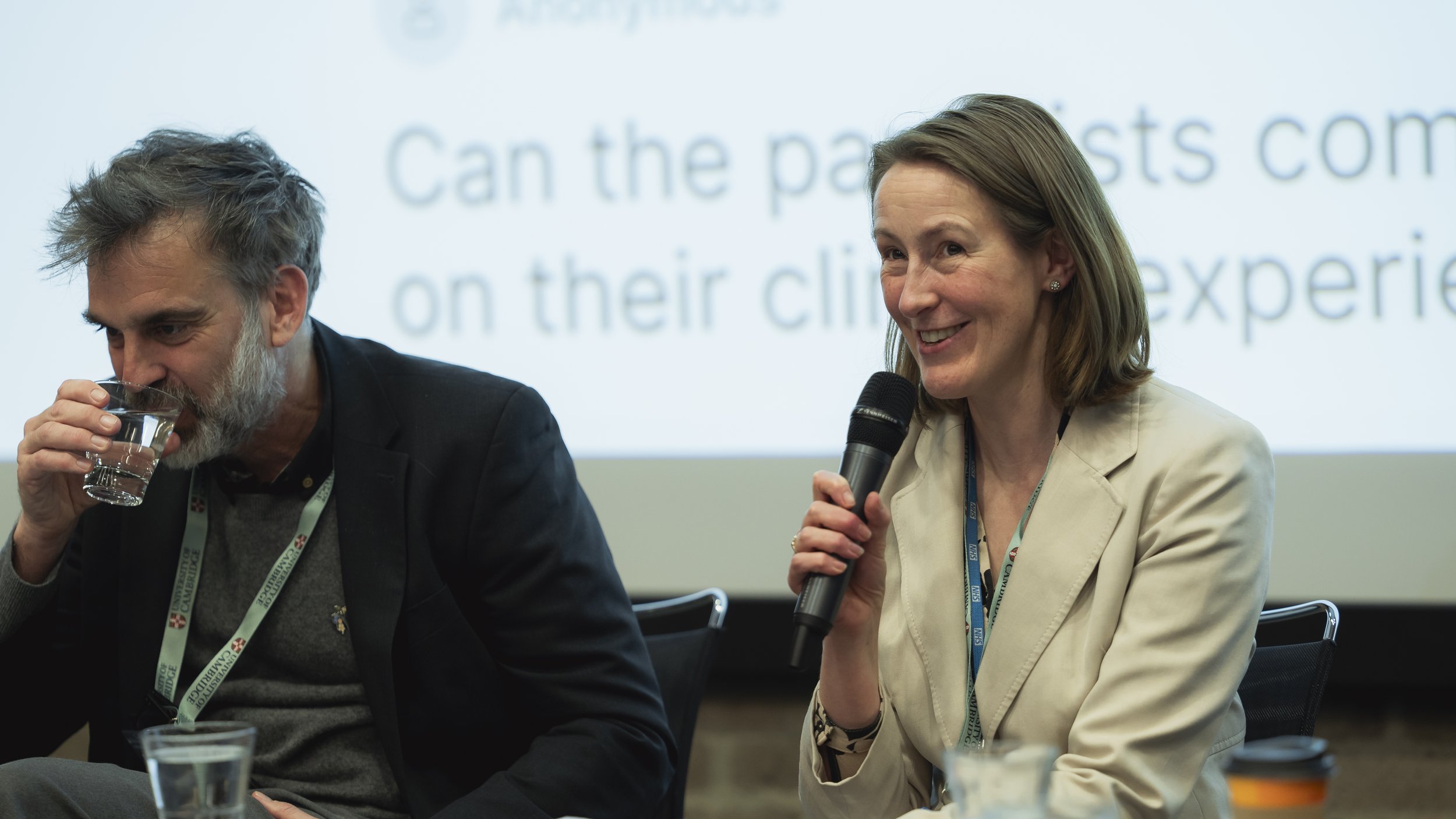 A woman smiling and speaking into a microphone during a panel discussion, with a man drinking water beside her, in front of a screen displaying partially visible text.