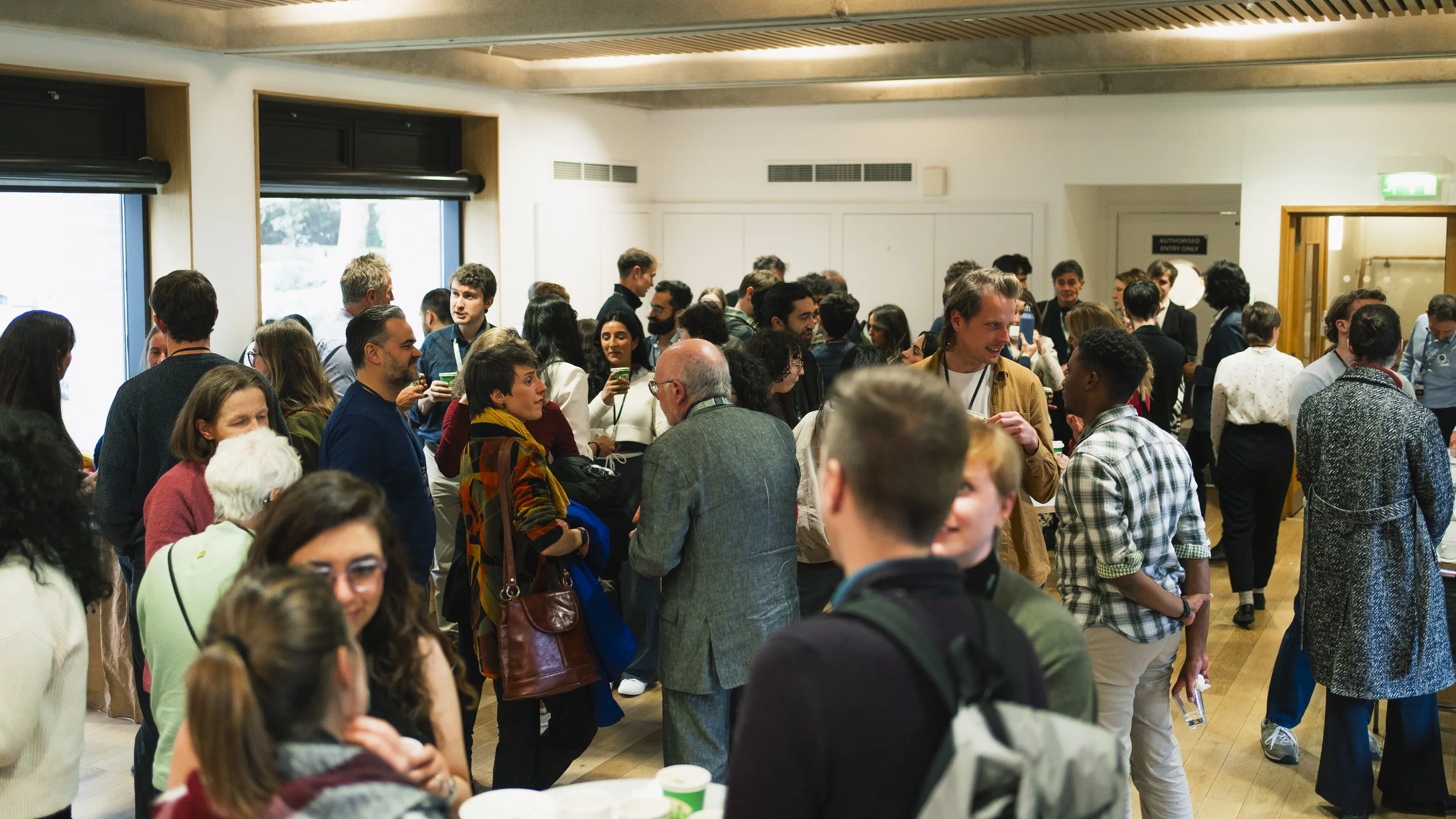 Crowd of diverse people at a social gathering or conference indoors with large windows and wooden decor, engaging in conversations.