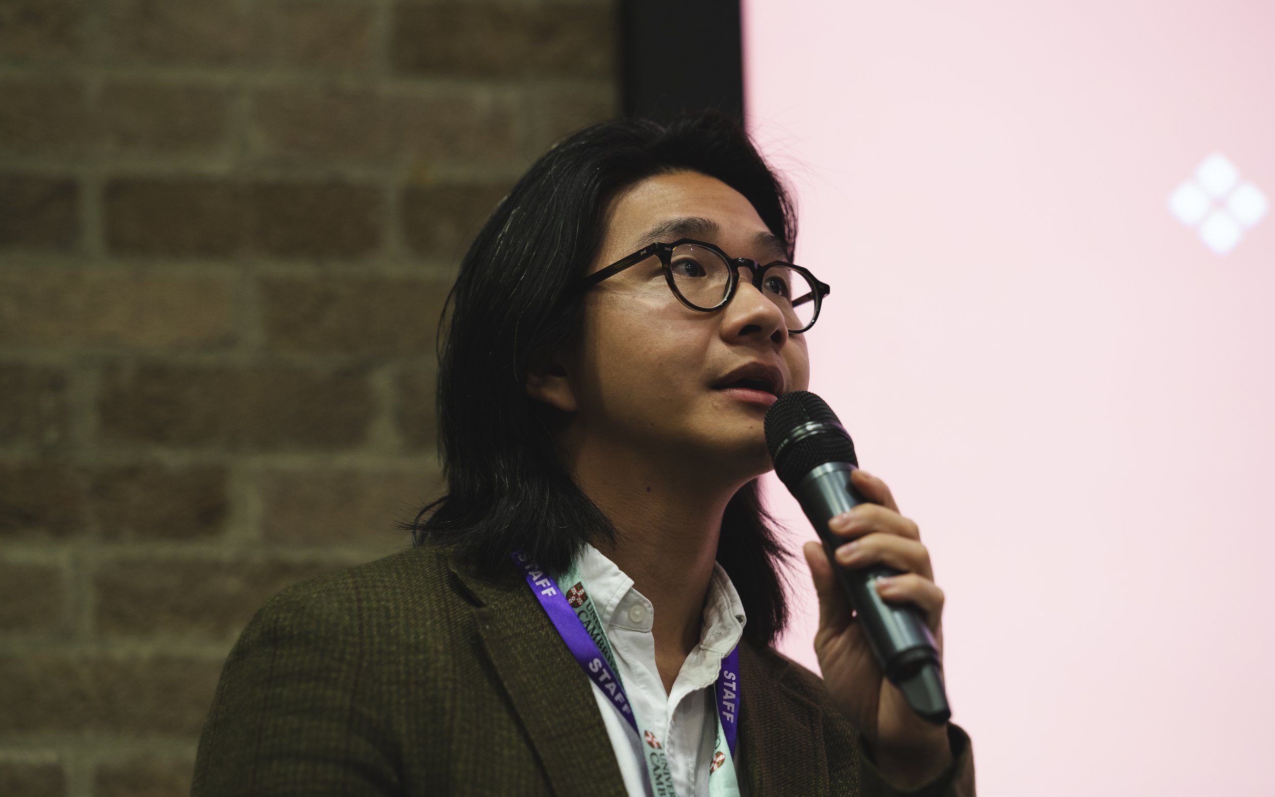 Woman with black hair and glasses speaking into a microphone during a presentation, with a brick wall on her left and a pink background on her right.