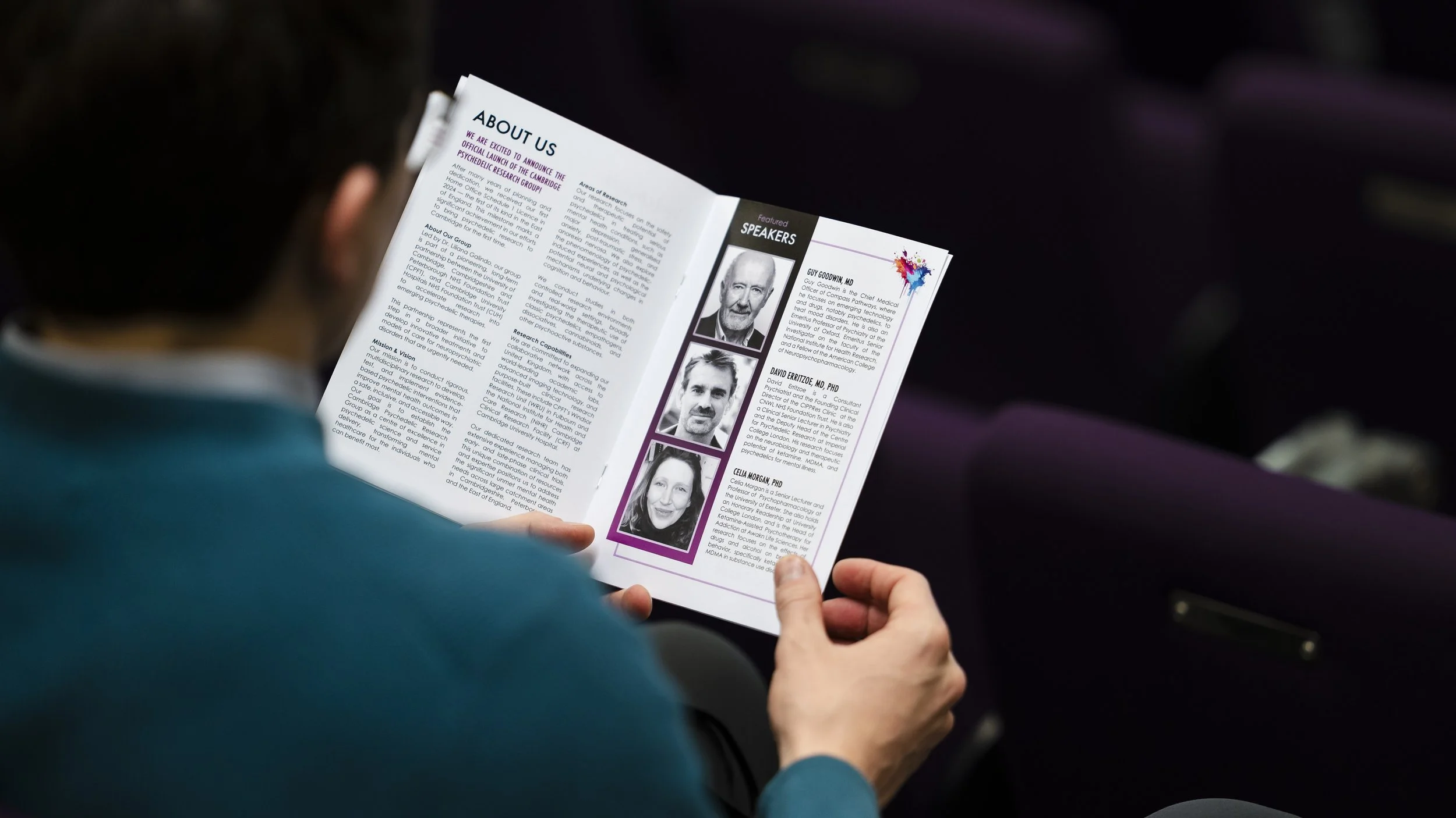 Person holding a brochure or magazine open to an 'About Us' page, which includes profiles of featured speakers.