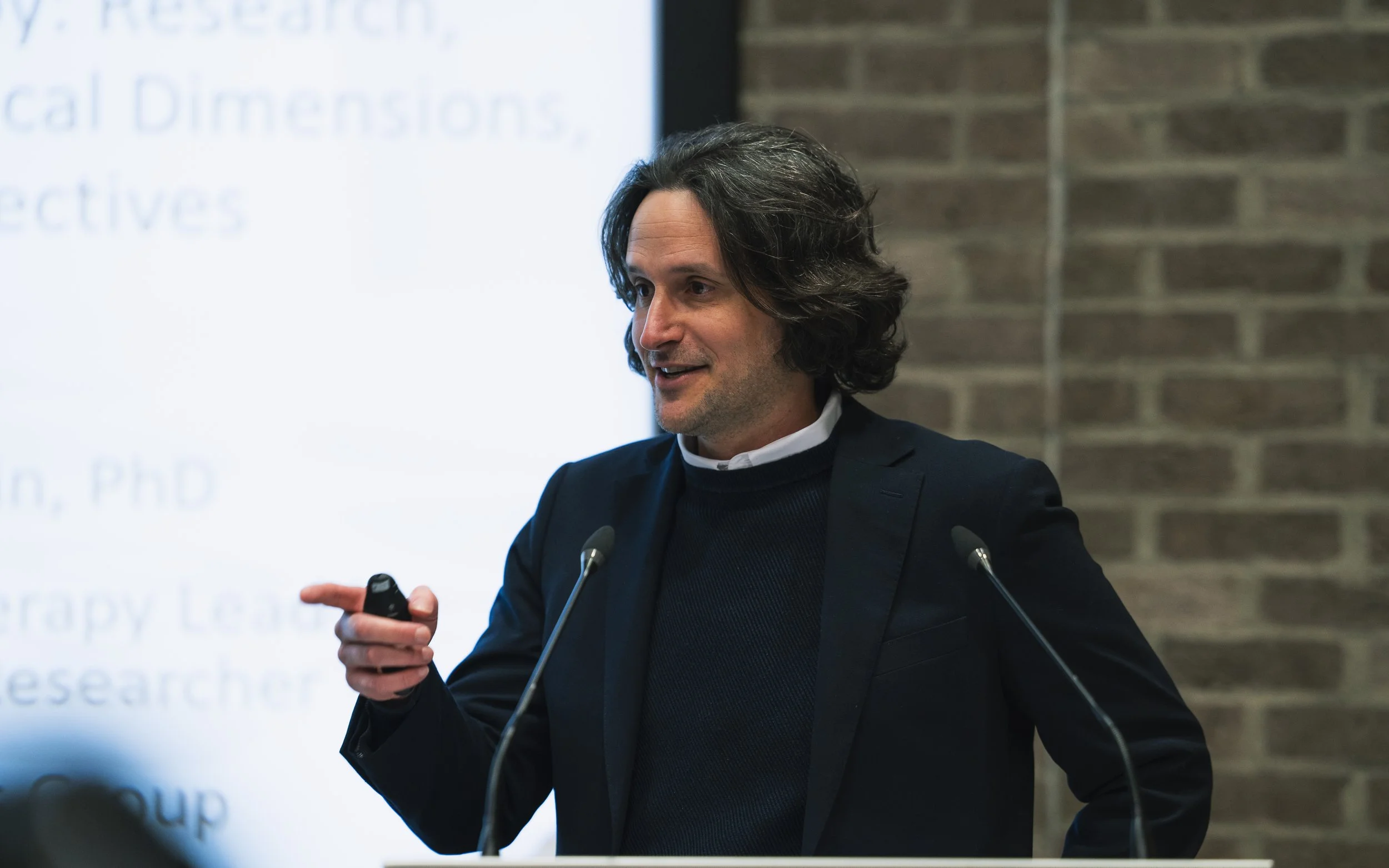 A man with shoulder-length dark hair wearing a dark blazer and shirt, speaking at a presentation behind a microphone, in front of a blurred presentation slide and brick wall.