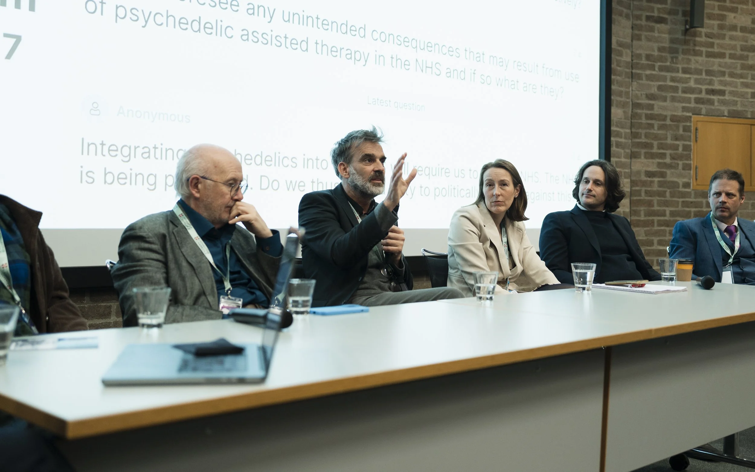 Panel of six people sitting at a long table during a conference or discussion, with a large screen displaying text behind them. The man in the middle is speaking and gesturing with his hand.