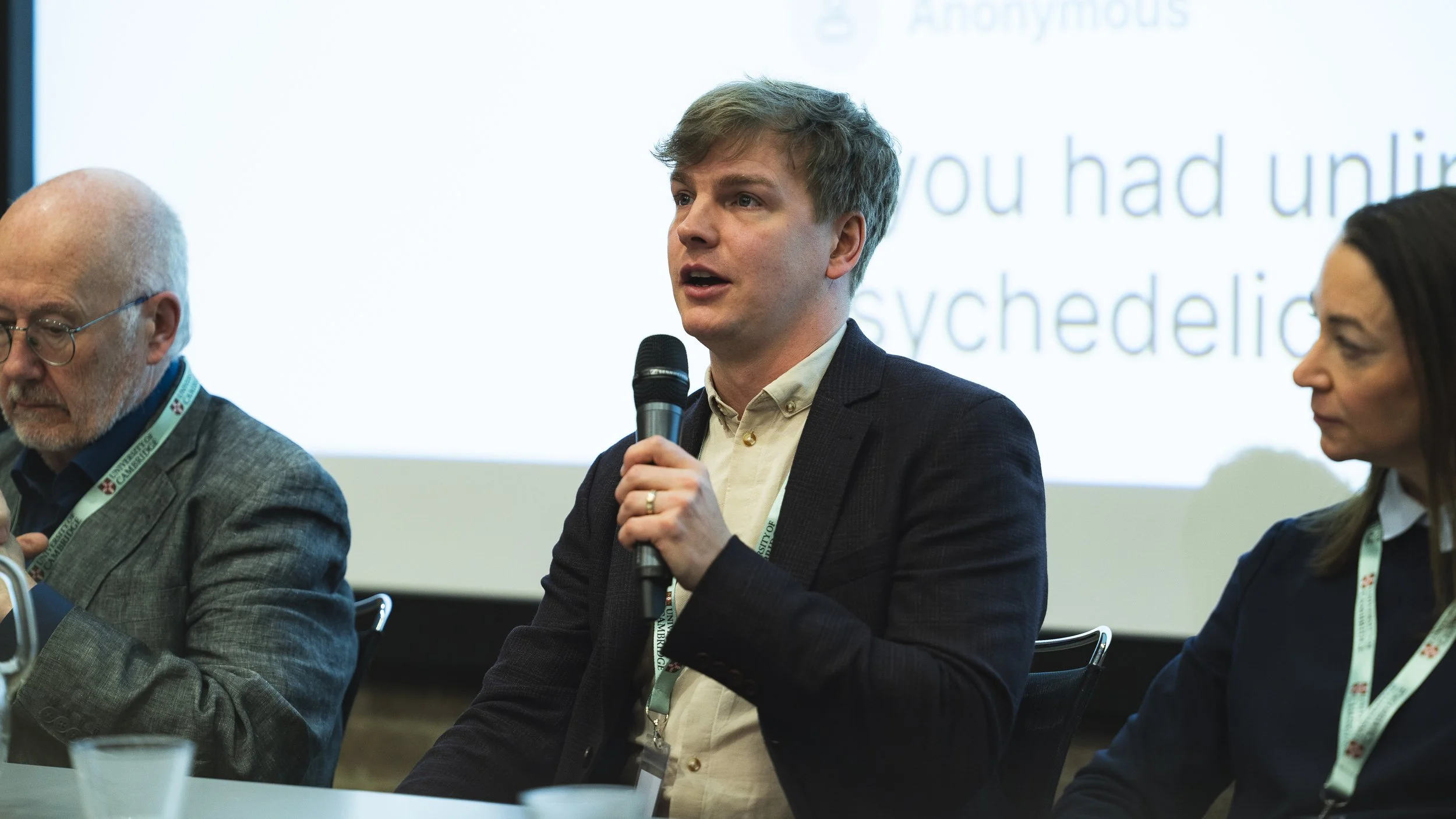 A man with light brown hair speaking into a microphone at a panel discussion, seated next to an elderly man with glasses on, at a conference or seminar.