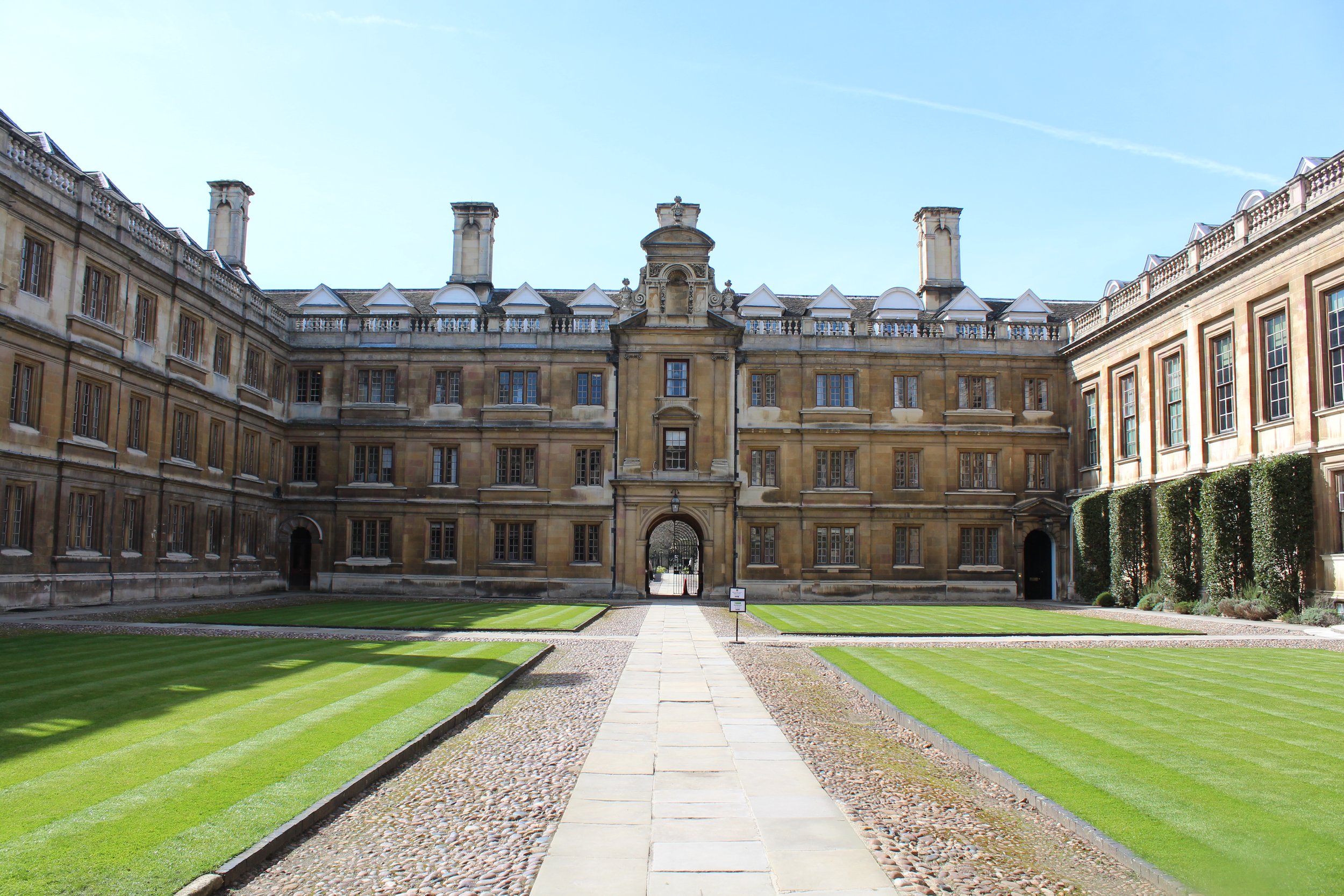 Courtyard with neatly manicured green grass, paved walkway, and historic stone building in the background under a clear blue sky.