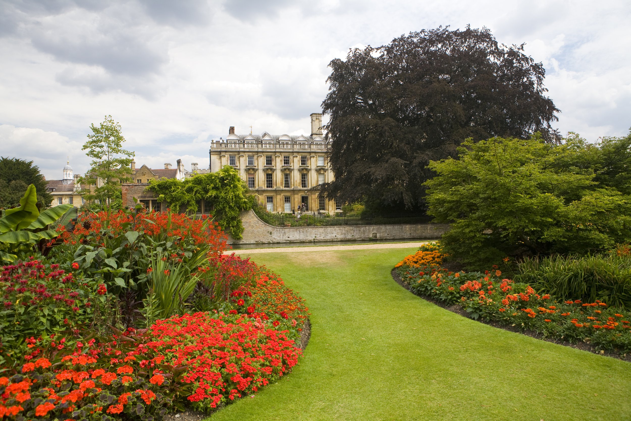 A historic building with large windows and a stone facade surrounded by a lush garden with colorful flowers, green shrubs, and tall trees, under a partly cloudy sky.
