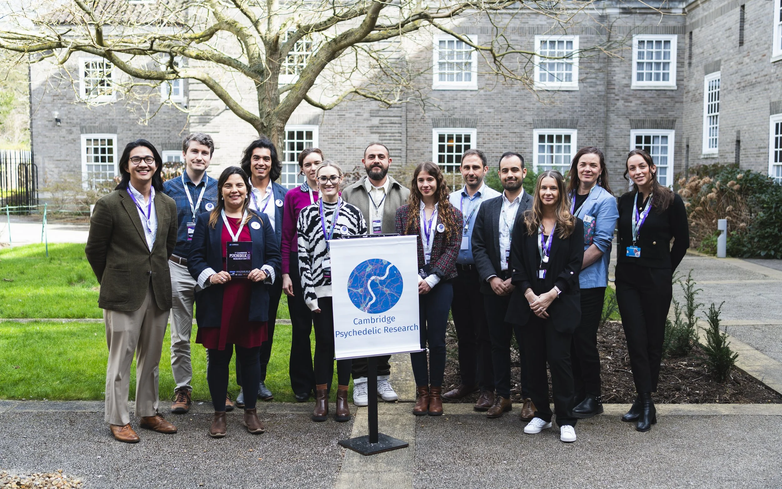 Group of people standing outdoors in front of a brick building, holding a sign that reads "Cambridge Psychedelic Research."