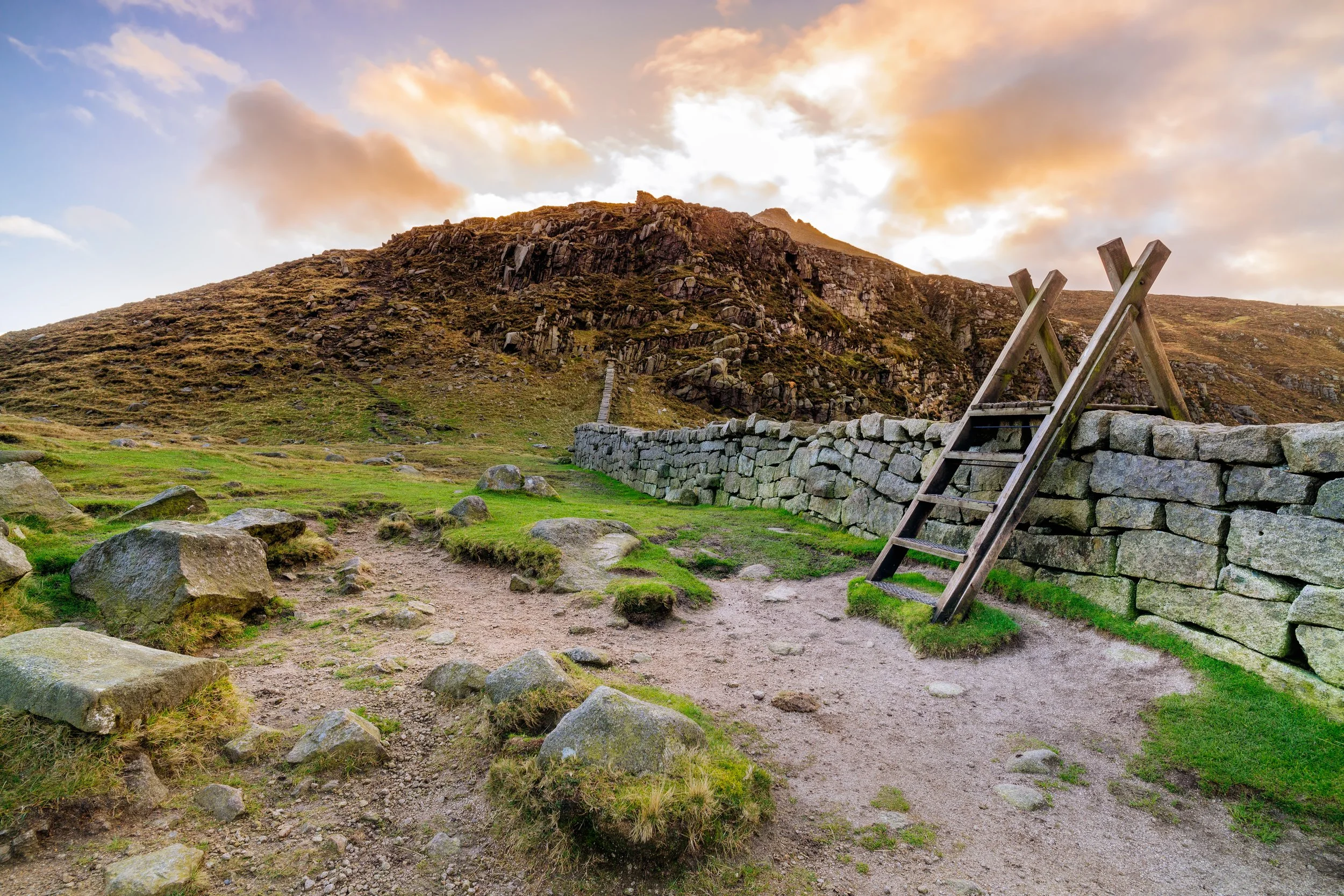 Stone wall with stile in a scenic hilly landscape at sunset, featuring grass and rocks.