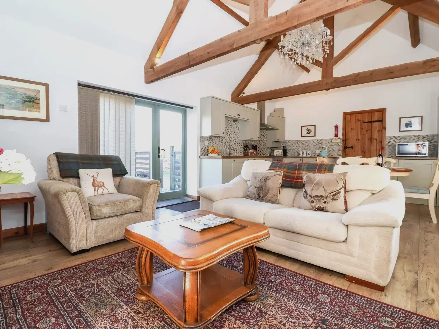 A cozy living room featuring a sofa with animal-themed pillows, a wooden coffee table, a armchair with a pillow, wooden beams, a fully kitted out kitchen with all you'll need.