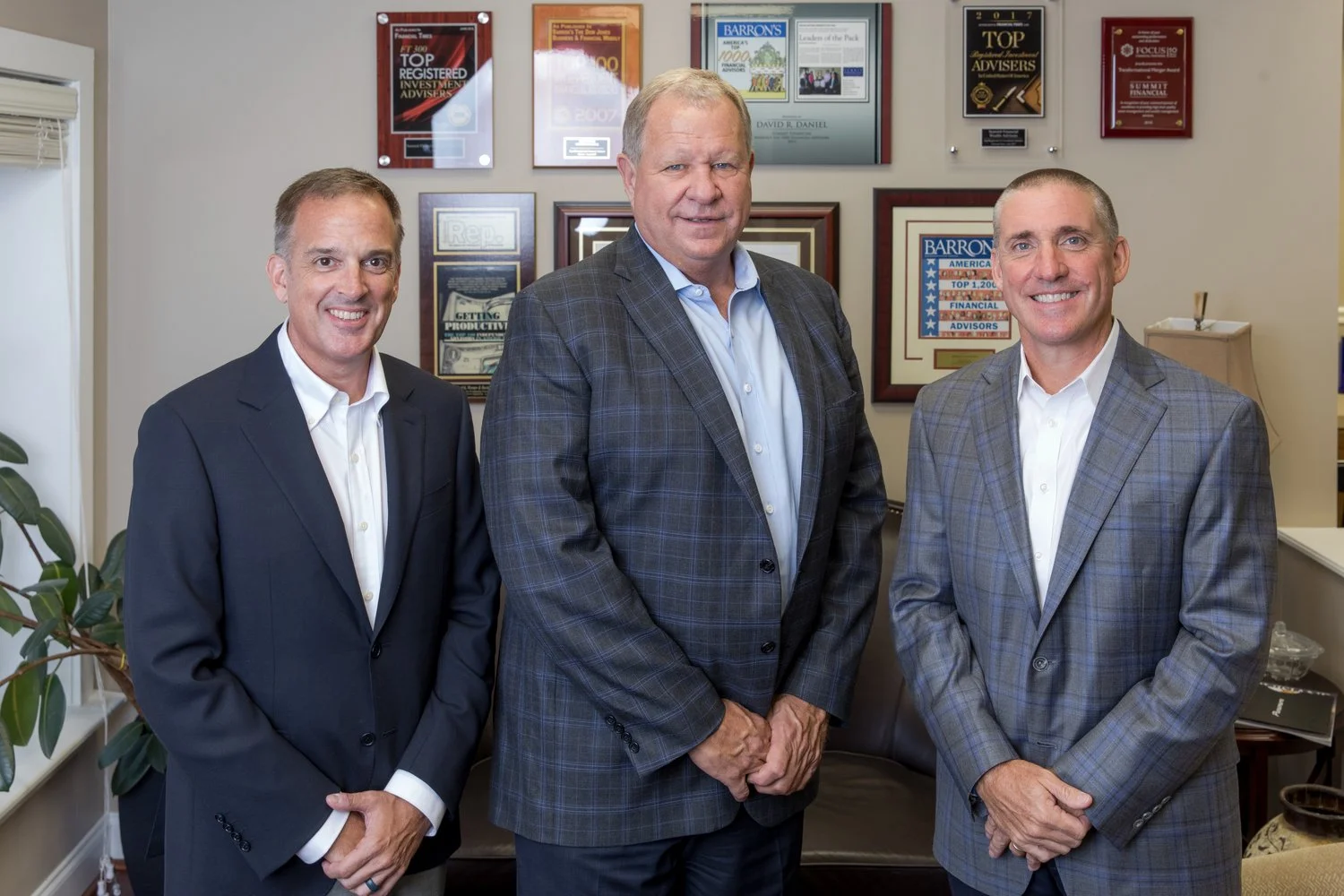Three men in suits standing in an office with award plaques and certificates on the wall behind them.