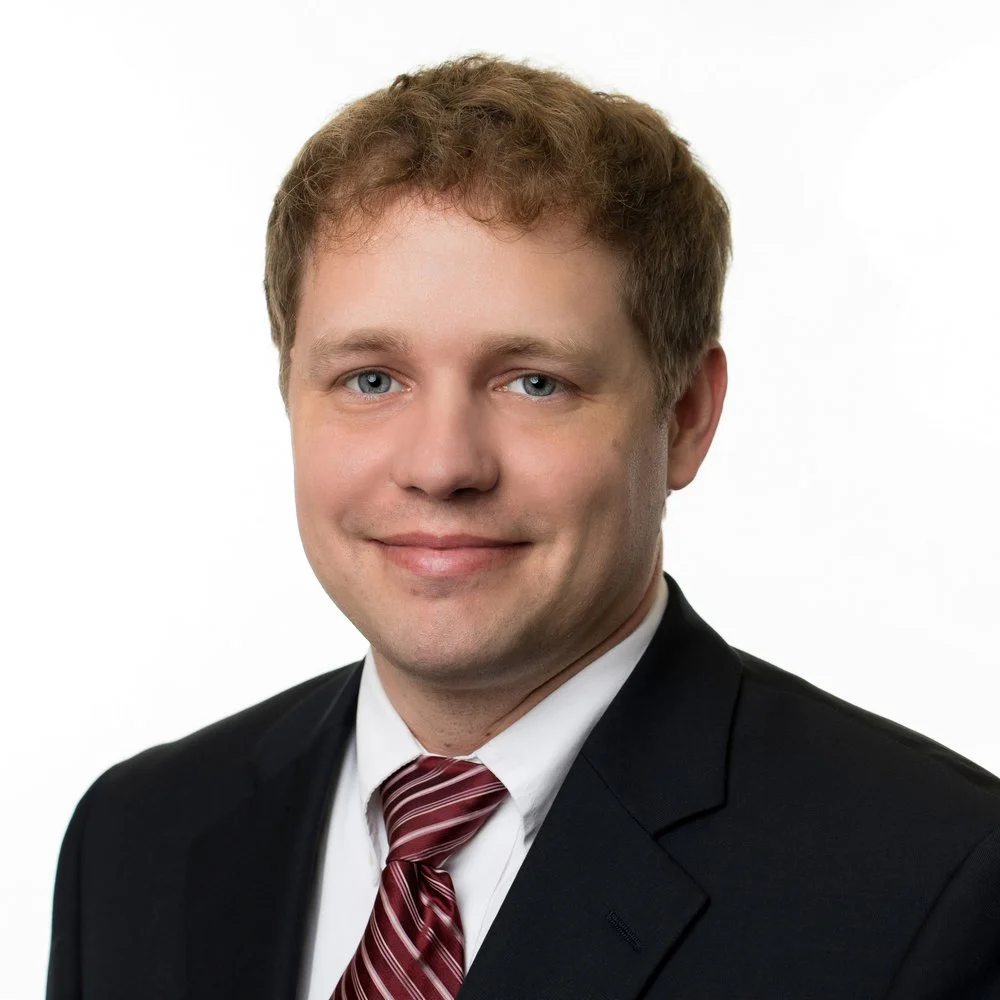 A professional headshot of a man with light brown hair, blue eyes, wearing a black suit, white shirt, and a red striped tie, smiling against a white background.