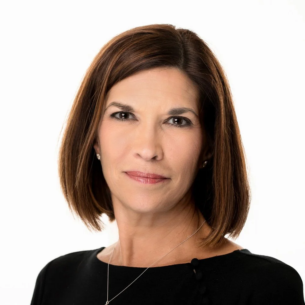 A woman with shoulder-length brown hair, wearing a black top and silver necklace, looking confidently at the camera against a plain white background.