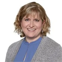 A smiling woman with curly blonde hair wearing a blue top and a gray cardigan, posed against a white background.