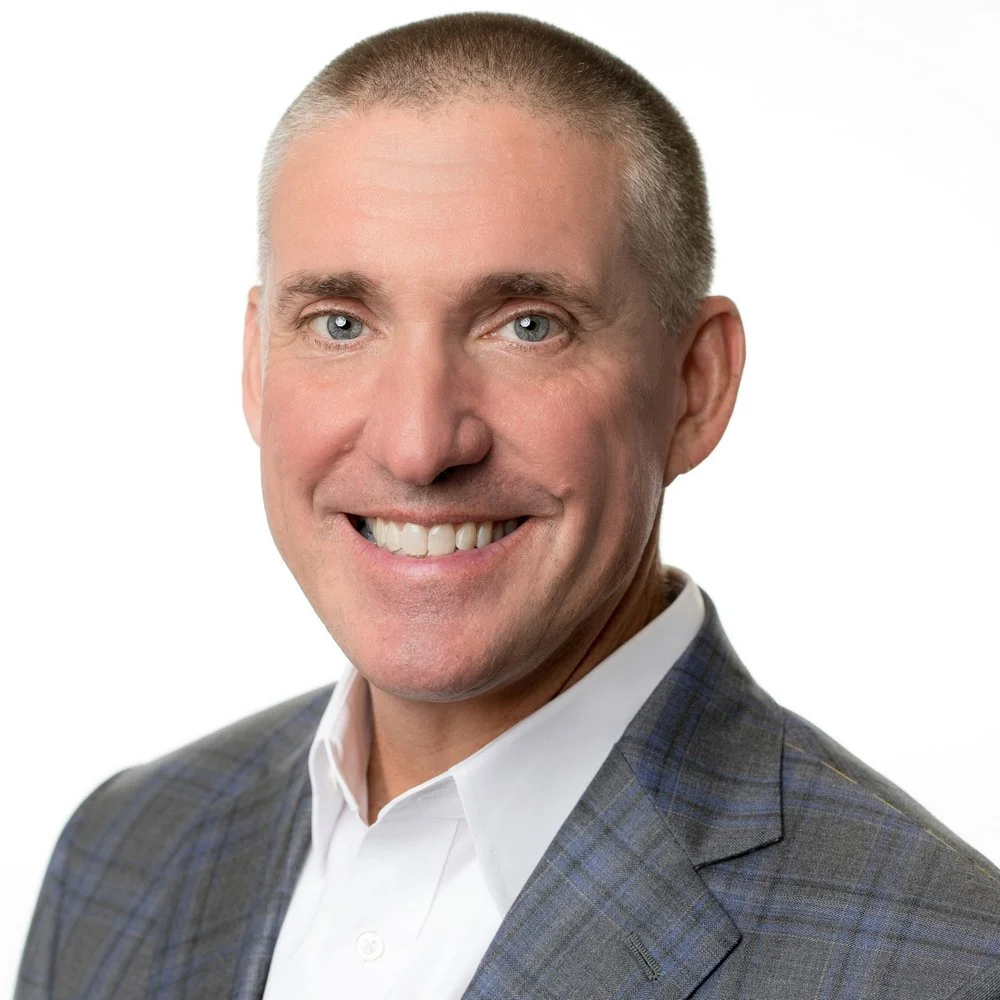 Portrait of a smiling man with short gray hair, wearing a white dress shirt and a gray plaid blazer, against a plain white background.