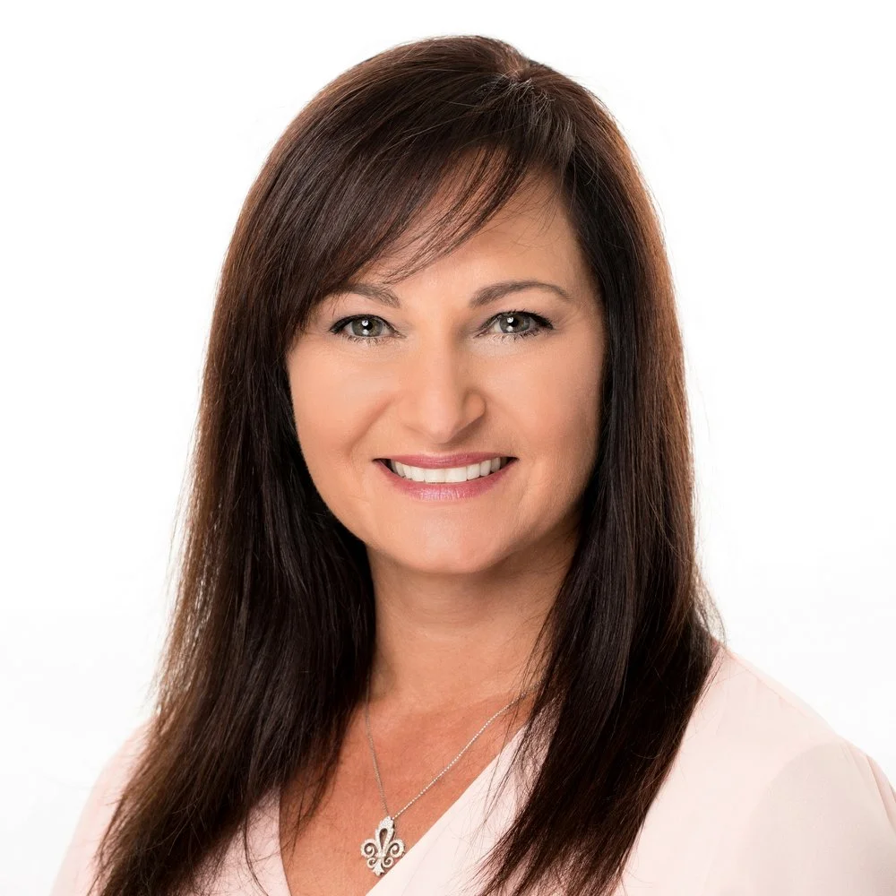A professional woman with long brown hair and a smile, wearing a light pink top and a silver necklace with a decorative pendant, against a white background.