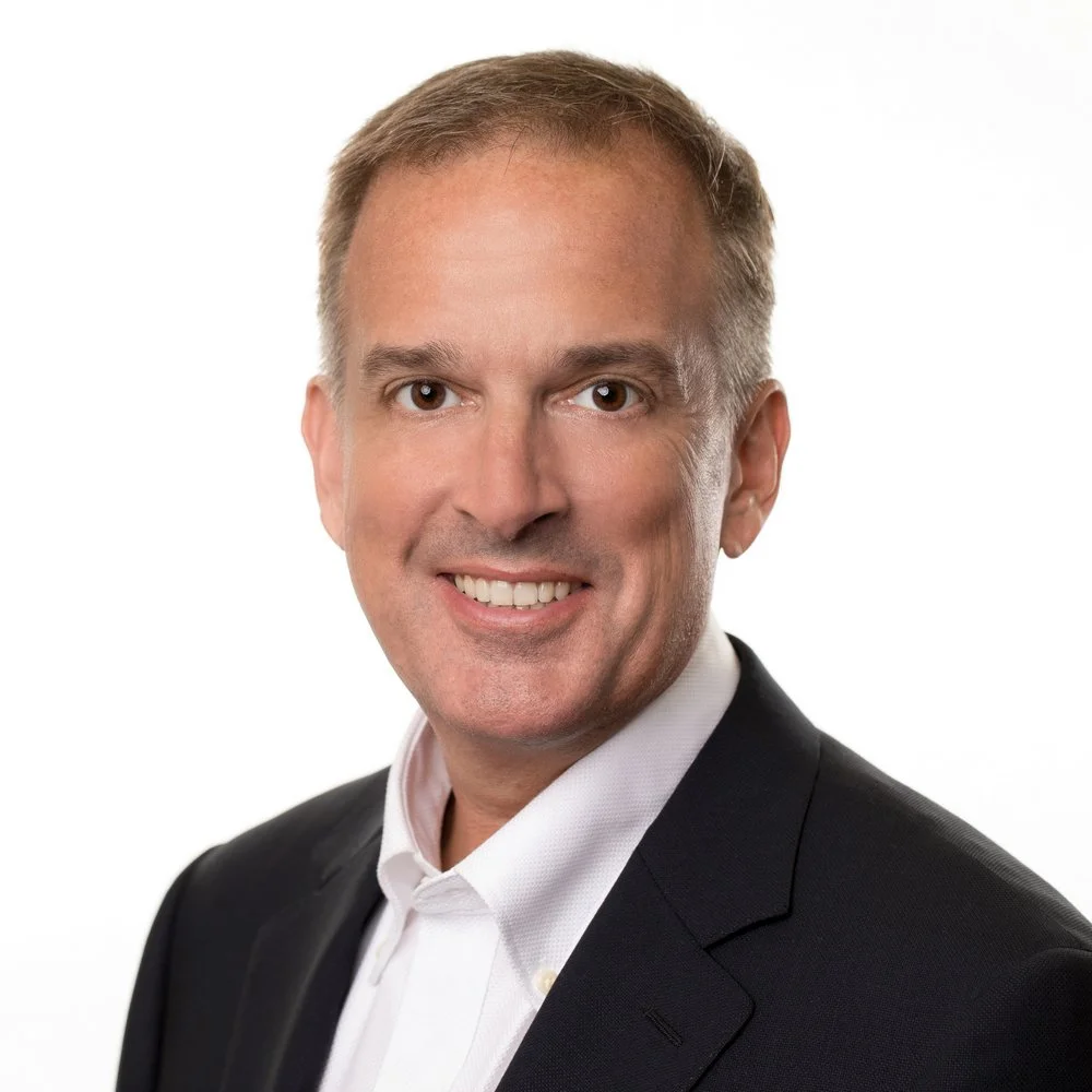 Professional headshot of a man with short blond hair, wearing a dark suit and white shirt, smiling against a white background.