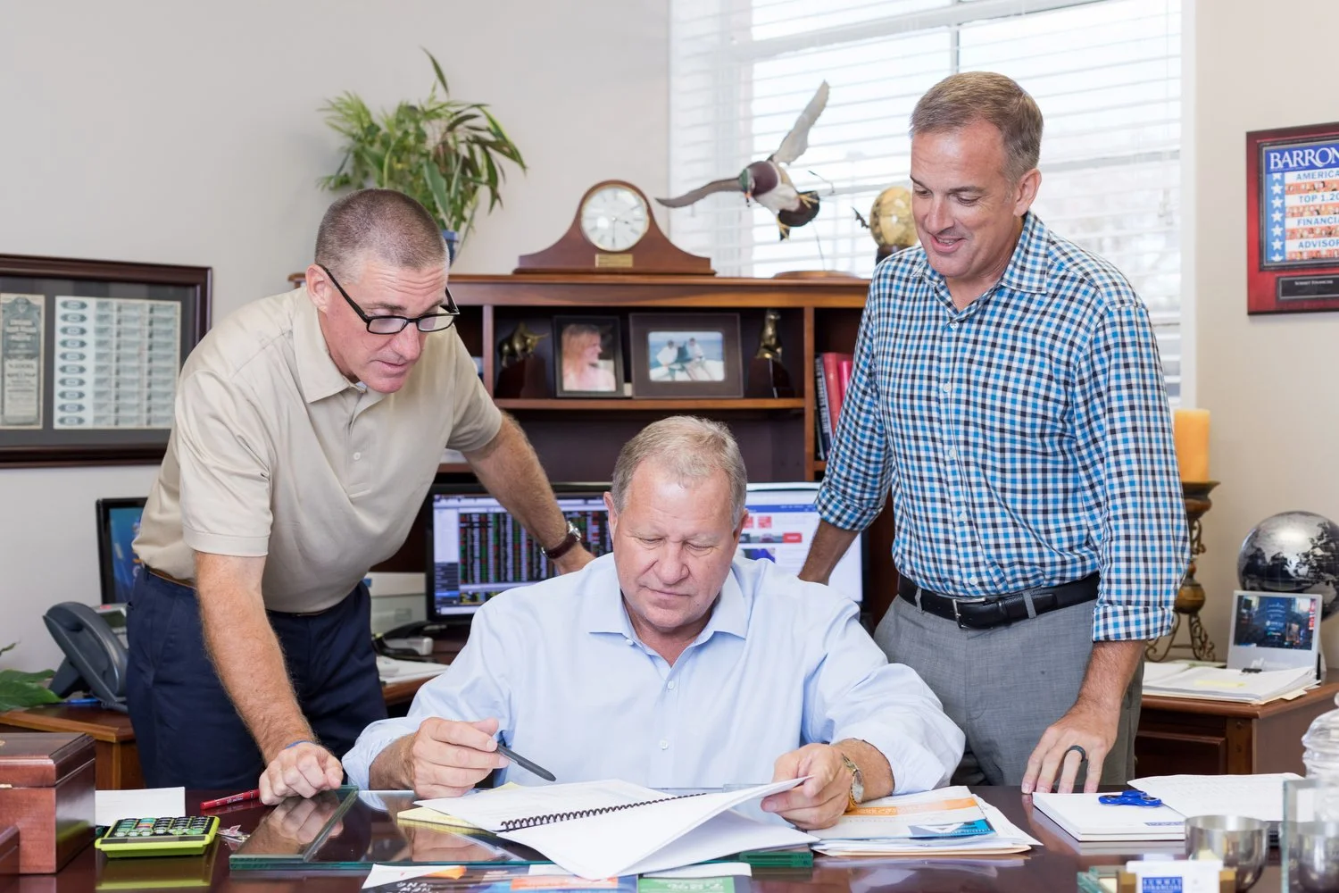 Three men in an office looking at documents on a desk, with one man seated and two men standing. The office has a wooden bookshelf with framed photos, a clock, and a model plane, a computer with financial graphs, and various office supplies.