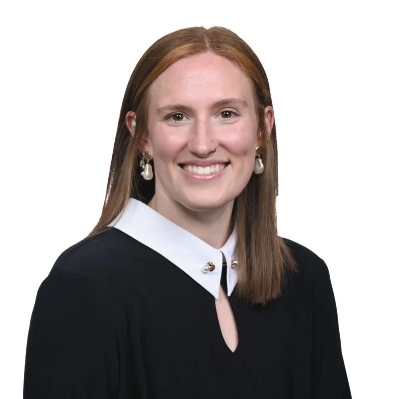 A young woman with straight red hair, light skin, and a bright smile, wearing pearl earrings, a black gown with a white collar, and standing against a plain white background.